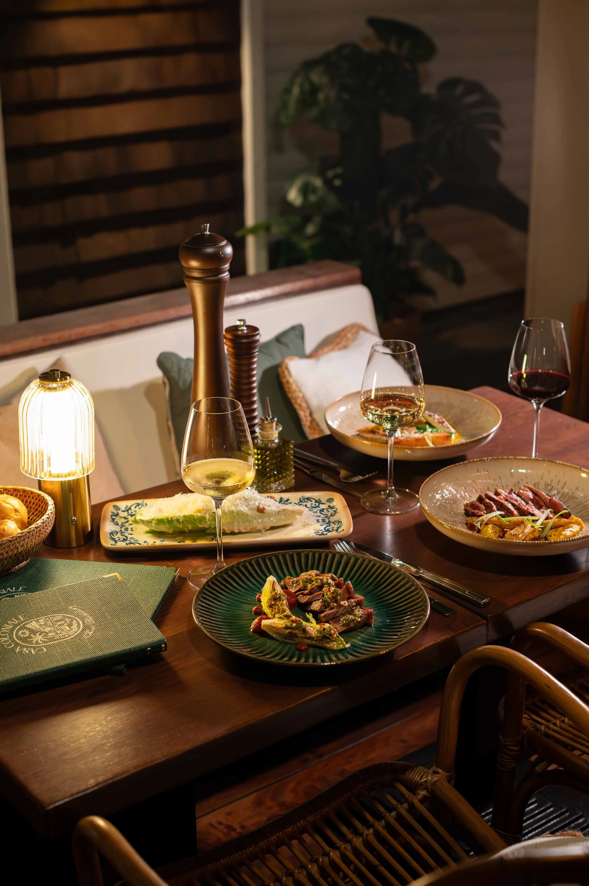 Table de dîner avec plusieurs plats, verres de vin blanc et rouge sous une lumière chaleureuse, décor intérieur avec plantes et lampes.