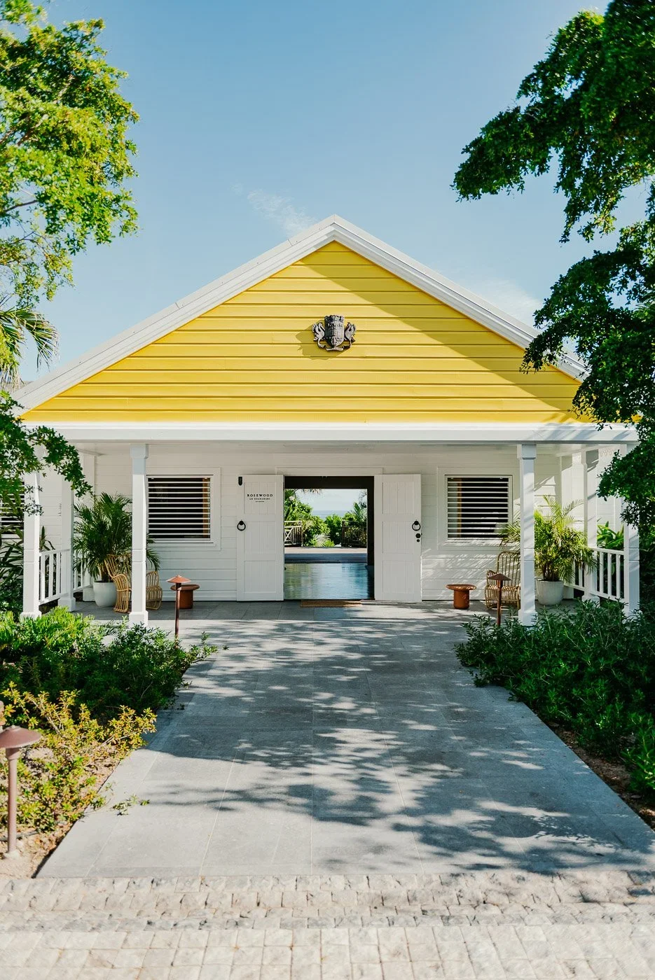 Maison en bois blanche avec un toit jaune, entourée d'arbres et de plantes, avec une allée en pierre devant l'entrée.