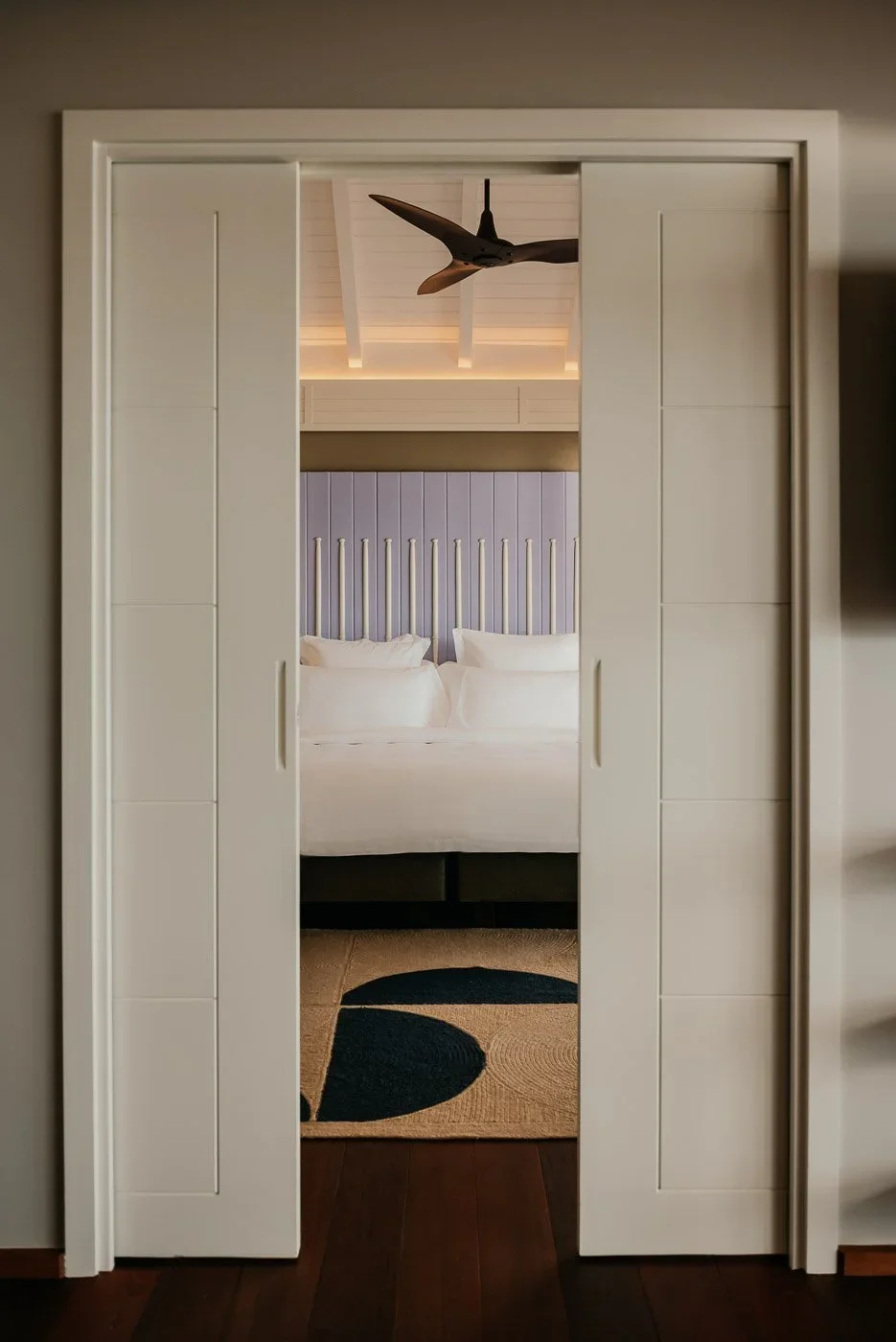 Une chambre vue à travers une porte coulissante blanche fermée, avec un lit blanc, une tête de lit en bois vertical, un plafond blanc avec une lampe et un ventilateur.