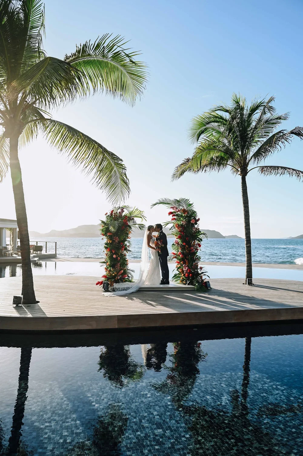 Un couple en mariage, debout sous une arche florale sur une plateforme en bois, près de la mer, avec des palmiers en arrière-plan et une piscine en avant-plan, lors d'une cérémonie de mariage au bord de l'eau.