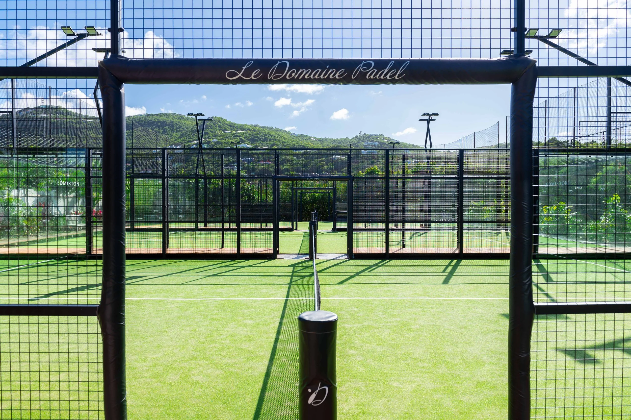 Terrain de padel entouré d'une clôture en métal, inscription 'Le Domaine Padel' en haut de l'entrée, avec un fond de collines verdoyantes et un ciel bleu avec quelques nuages.