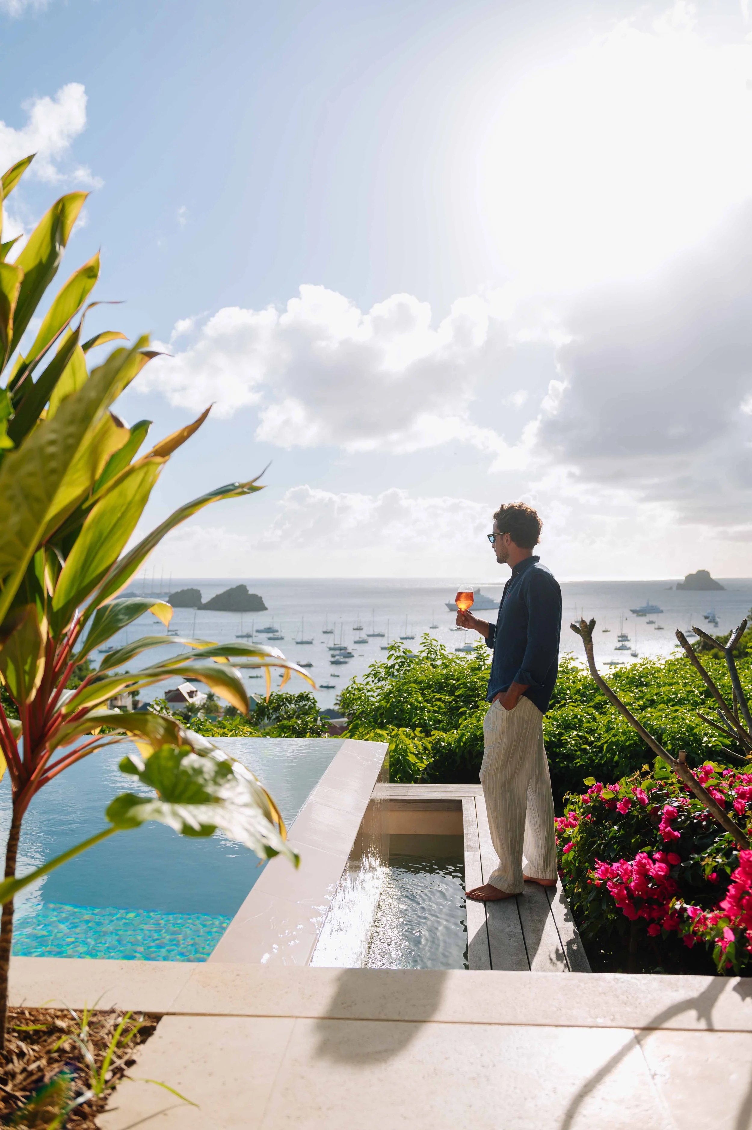 Un homme debout près d'une piscine, tenant un verre de boisson, avec une vue sur la mer et des yachts en arrière-plan, parmi des plantes et des fleurs colorées.