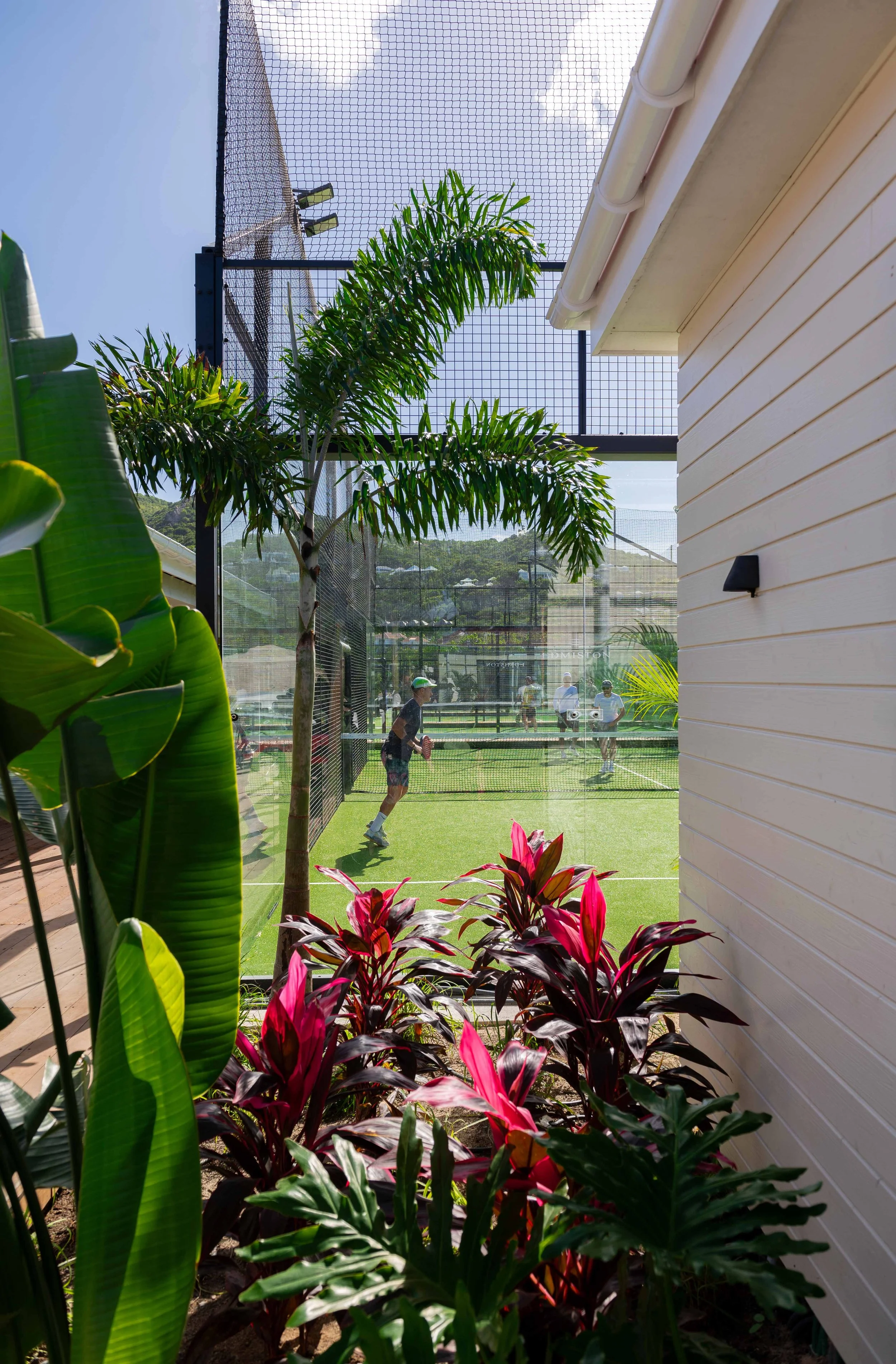 Terrasse avec des plantes vertes et un court de paddle tennis où des personnes jouent au tennis indoor.