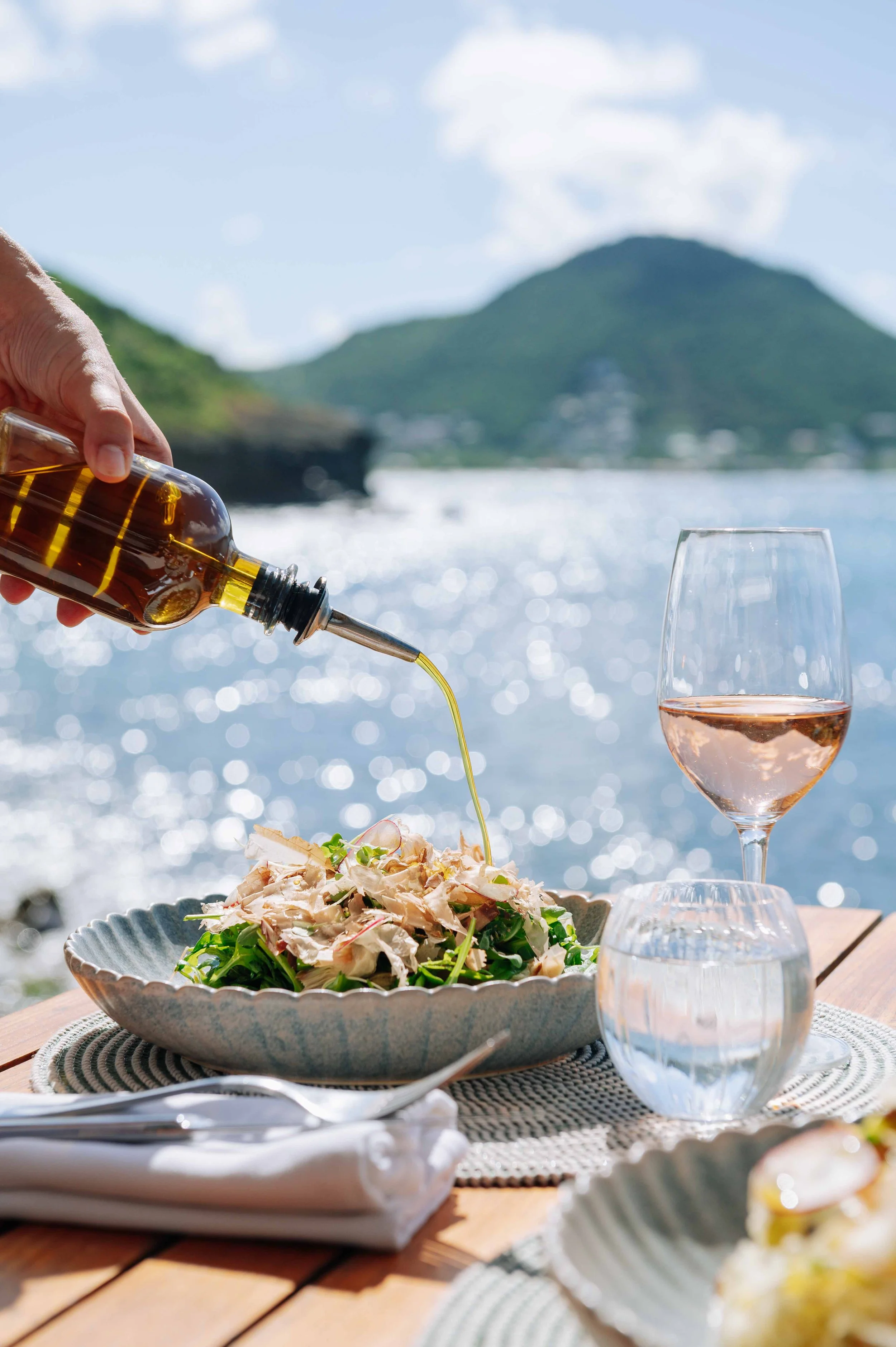 Une personne verse de l'huile d'olive sur une salade dans un décor en bord de mer avec des montagnes en arrière-plan, une verre de vin rosé et un verre d'eau sur une table en bois.