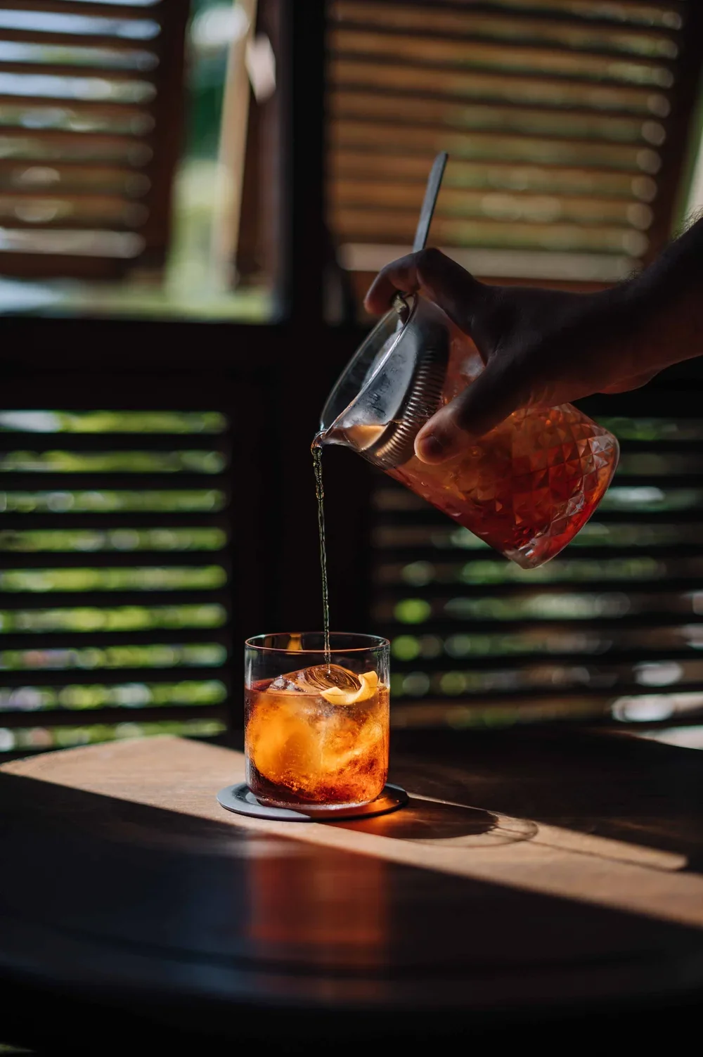 Une main verse un liquide brunâtre dans un verre contenant des glaçons et une tranche d'orange, sur une table en bois, avec un fond de fenêtres en lattes en bois.
