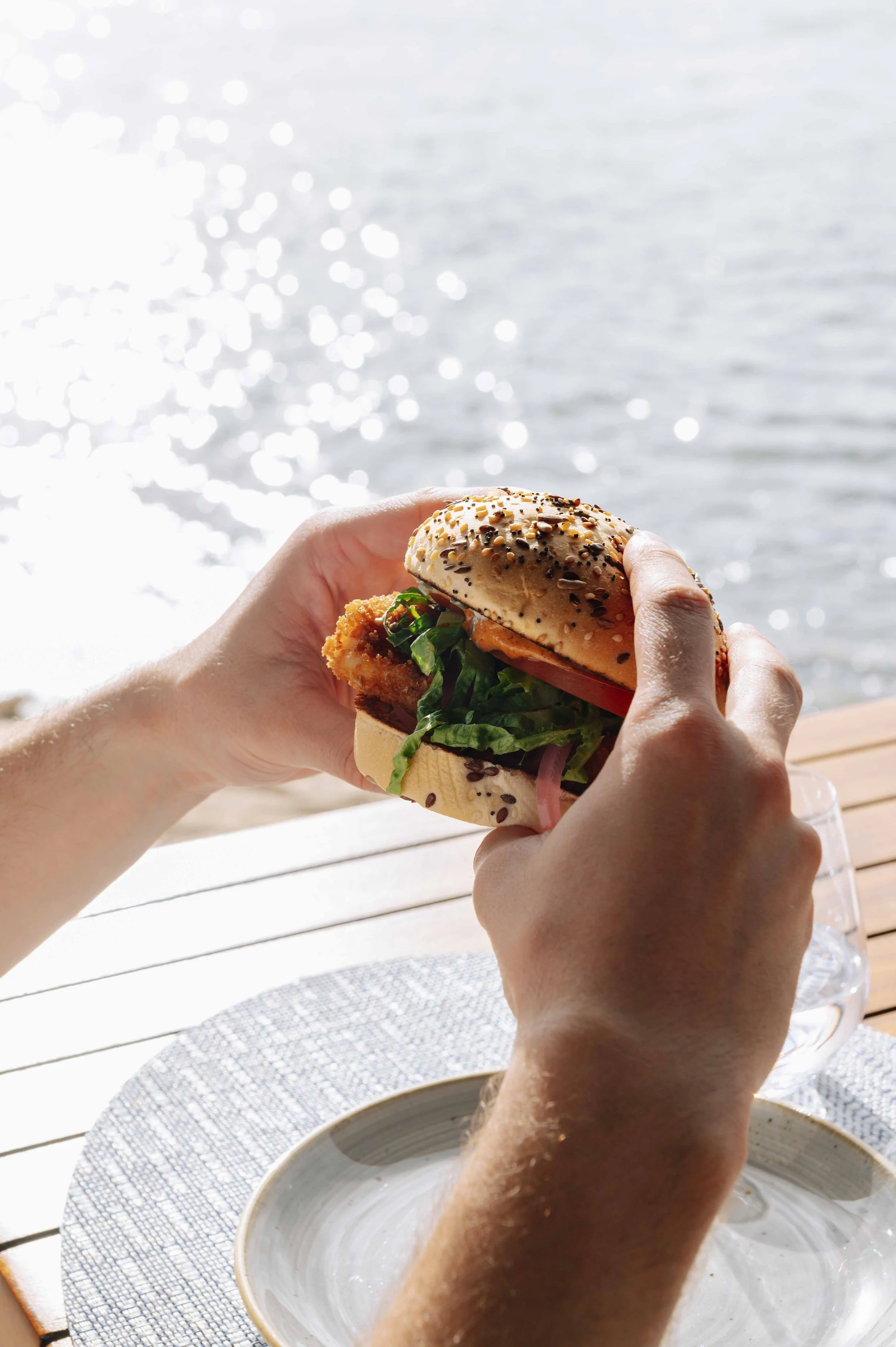 Person tenant un sandwich avec des légumes et du poisson, près d'une étendue d'eau ensoleillée.