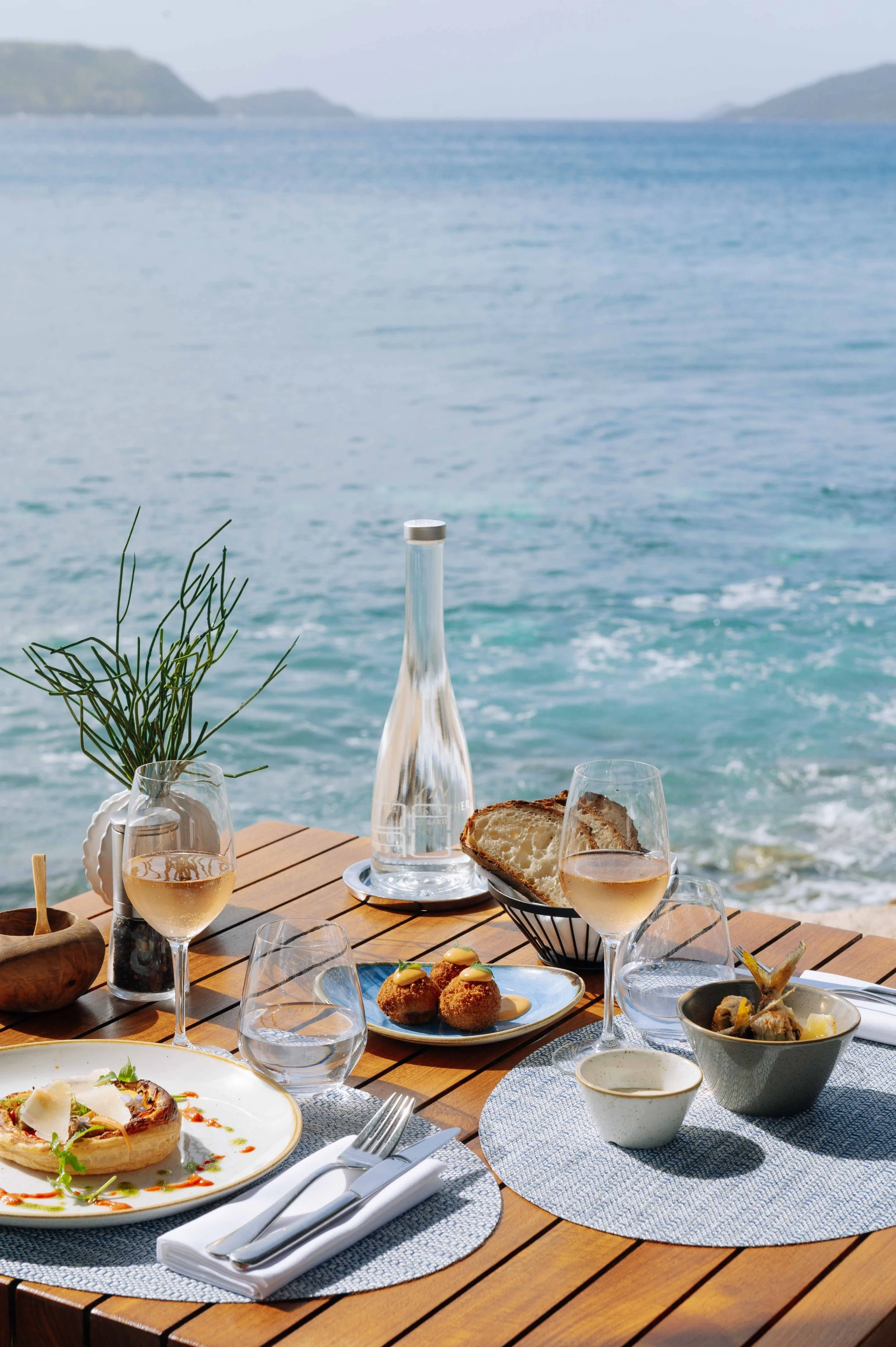Repas en plein air sur une terrasse en face de la mer avec des plats, verres de vin blanc, pain, et une bouteille d'eau.