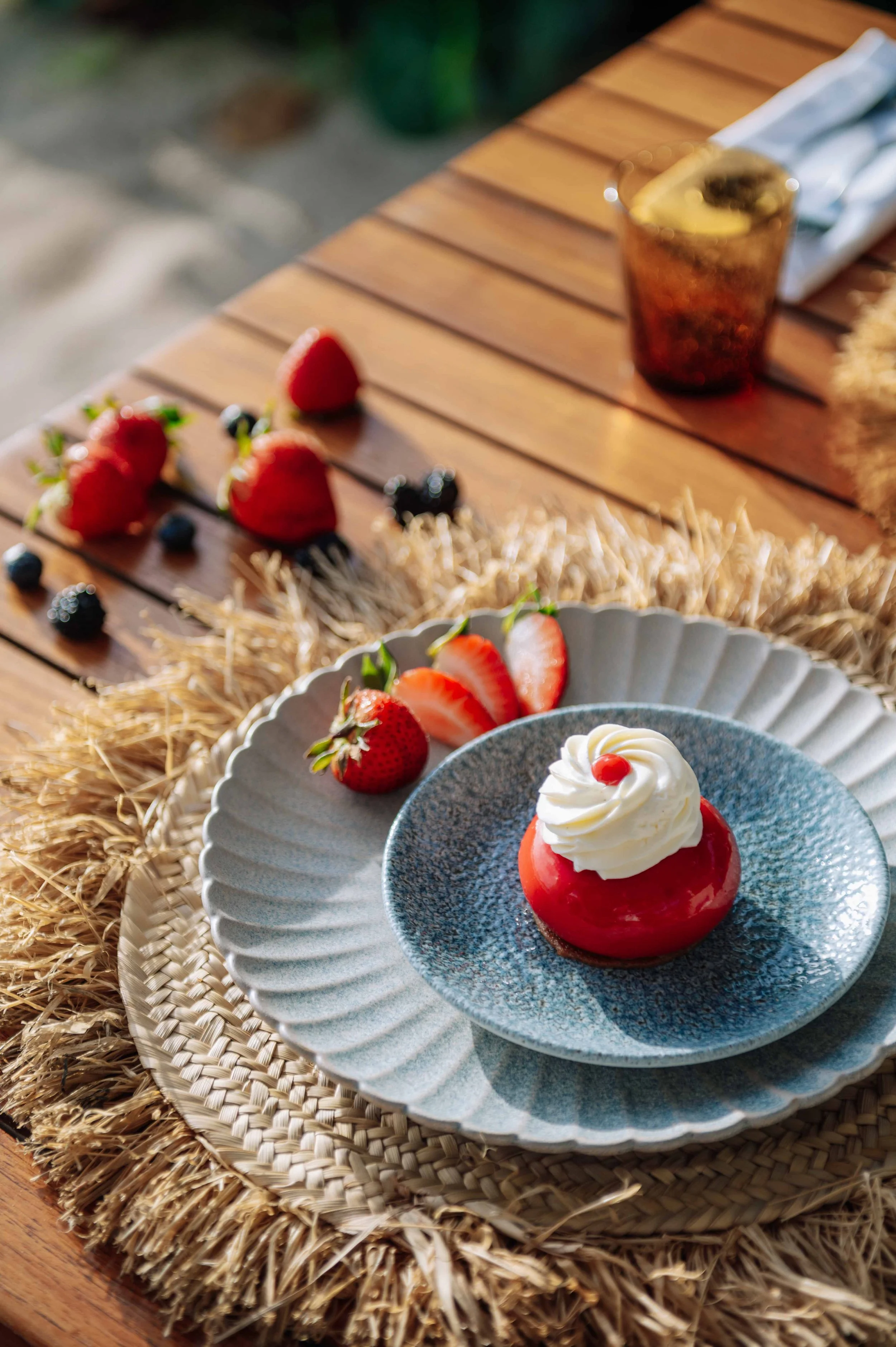 Un dessert à la fraise avec de la crème fouettée sur un plateau, entouré de fraises et de morceaux de fraises, avec d'autres fruits et une boisson en arrière-plan sur une table en bois.