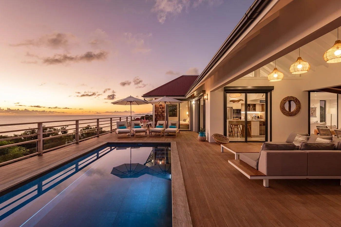 Terrasse avec piscine, chaises longues sous parasols, vue sur la mer au coucher du soleil, intérieur moderne avec canapé, lampes suspendues en rotin, cuisine ouverte.