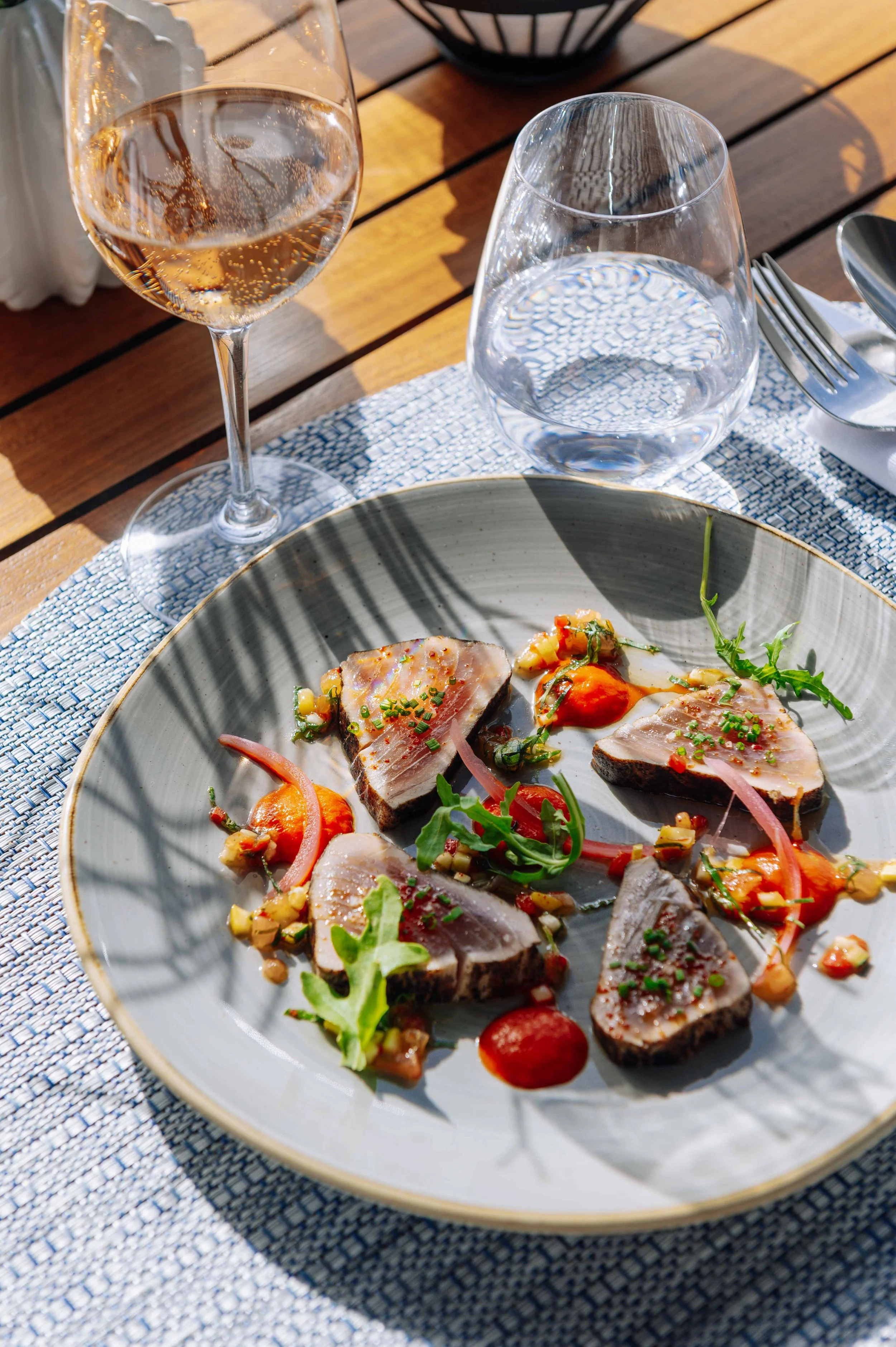 Assiette de tartare de poisson avec des garnitures et sauce, verre de vin blanc, verre d'eau, couverts, nappe en tissu sur une table en bois.