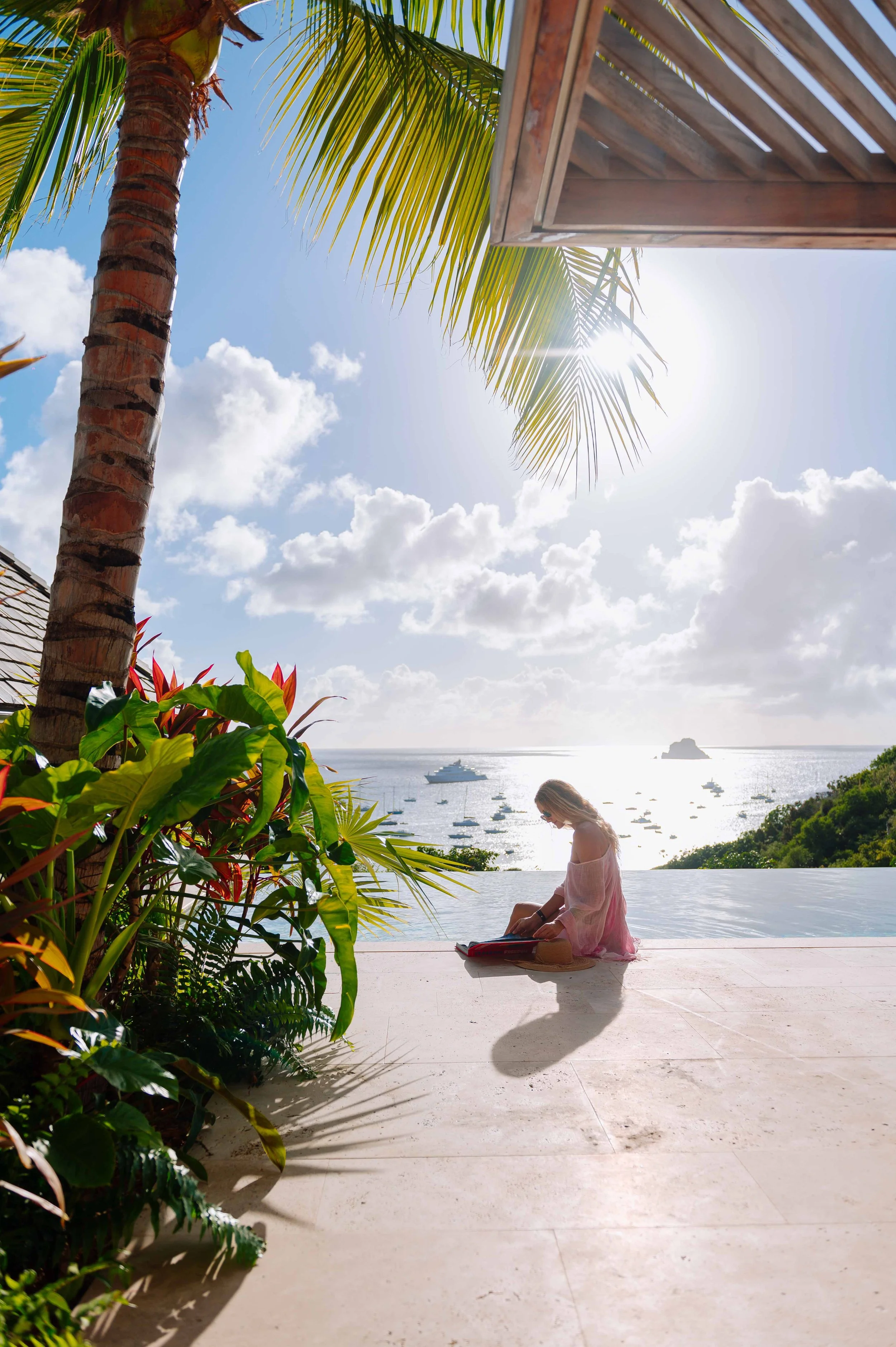 Une femme assise près d'une piscine en regardant un livre, avec la mer en arrière-plan, entourée de végétation tropicale et sous un ciel ensoleillé.