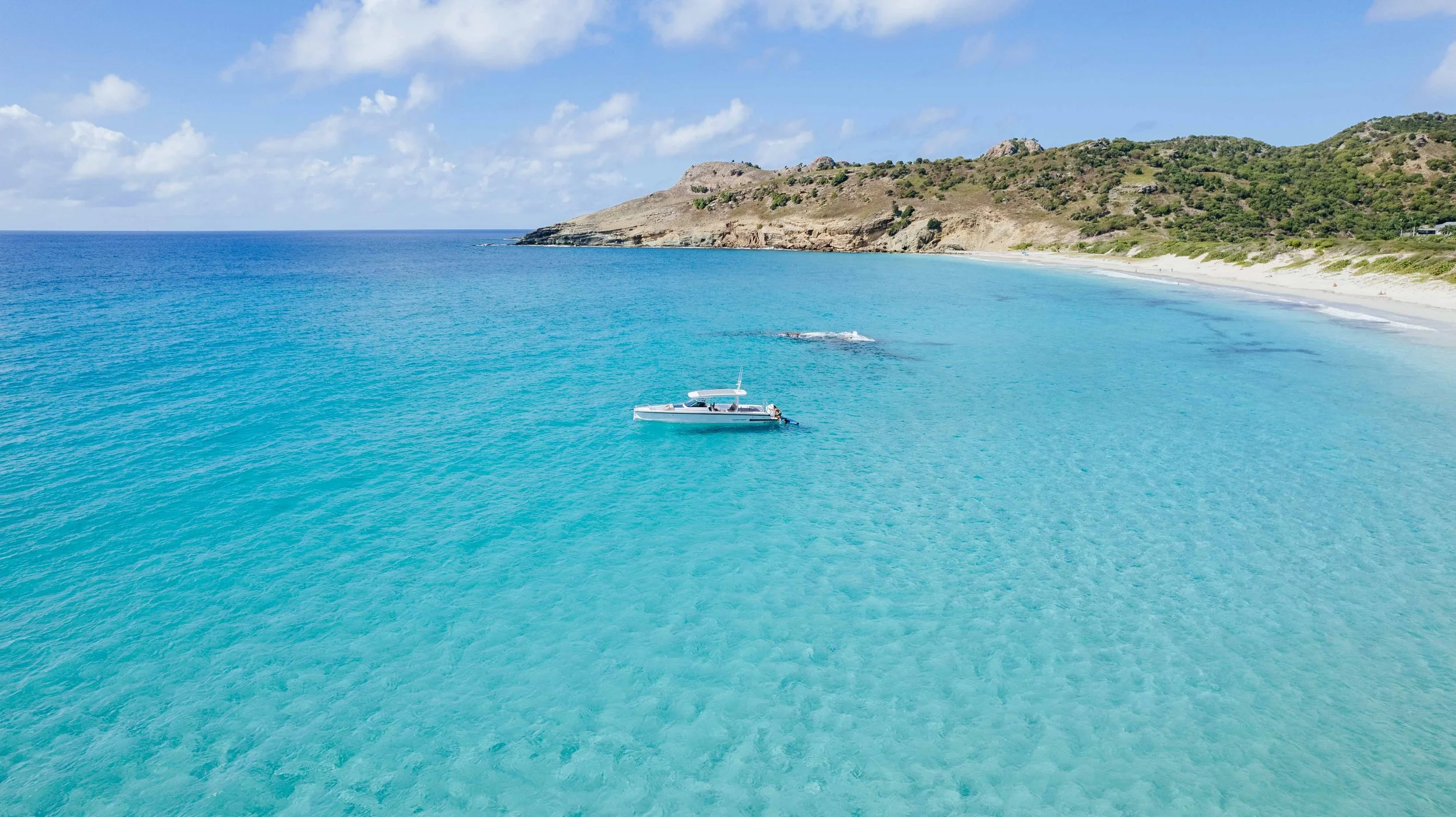 Vue aérienne d'une plage de sable blanc avec une mer turquoise, un bateau à moteur flottant à proximité, escarpés avec de la végétation en arrière-plan, ciel ensoleillé avec quelques nuages.