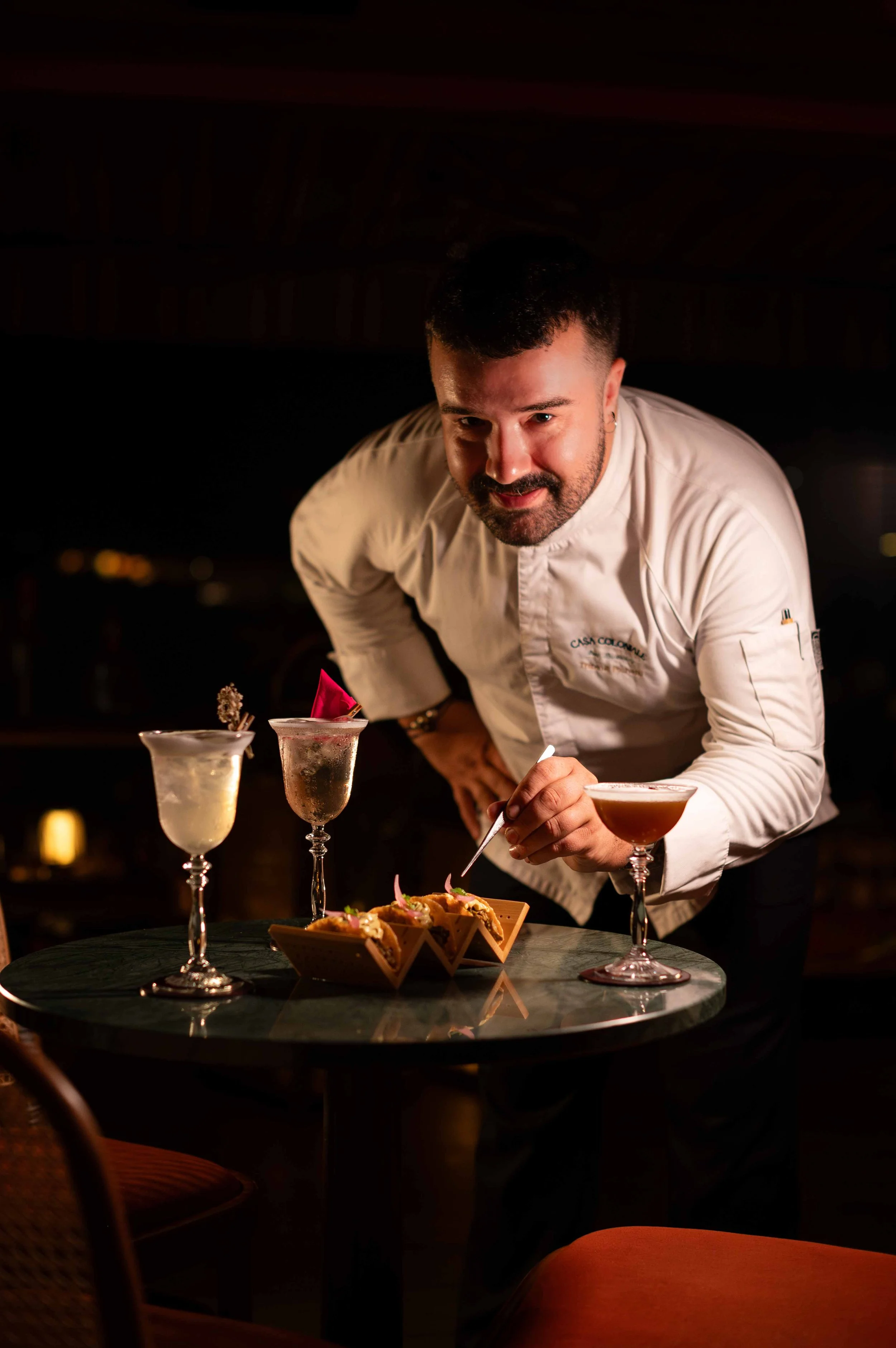 Un chef en tenue blanche prépare des amuse-bouches dans un restaurant élégant la nuit, avec des cocktails sur la table.