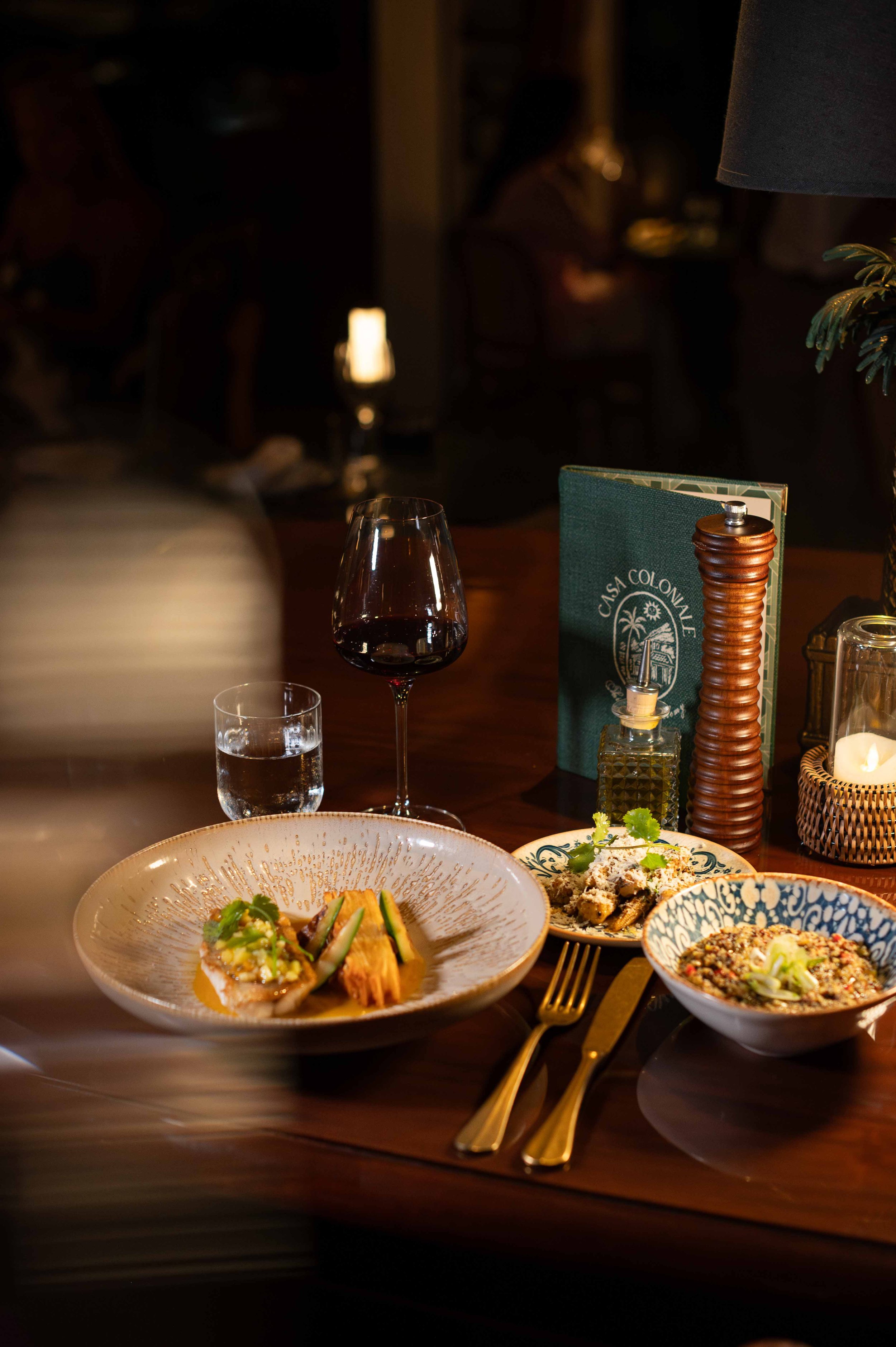 Table avec trois plats gastronomiques, un verre de vin rouge, un verre d'eau, une bouteille d'huile, un menu, une bougie, et des accessoires de table.