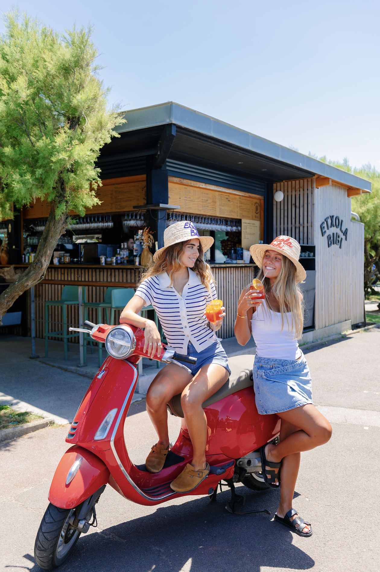Two women sitting on a red scooter outside a cafe, wearing summer dresses and straw hats, holding drinks, smiling, with a tree and a cafe in the background.