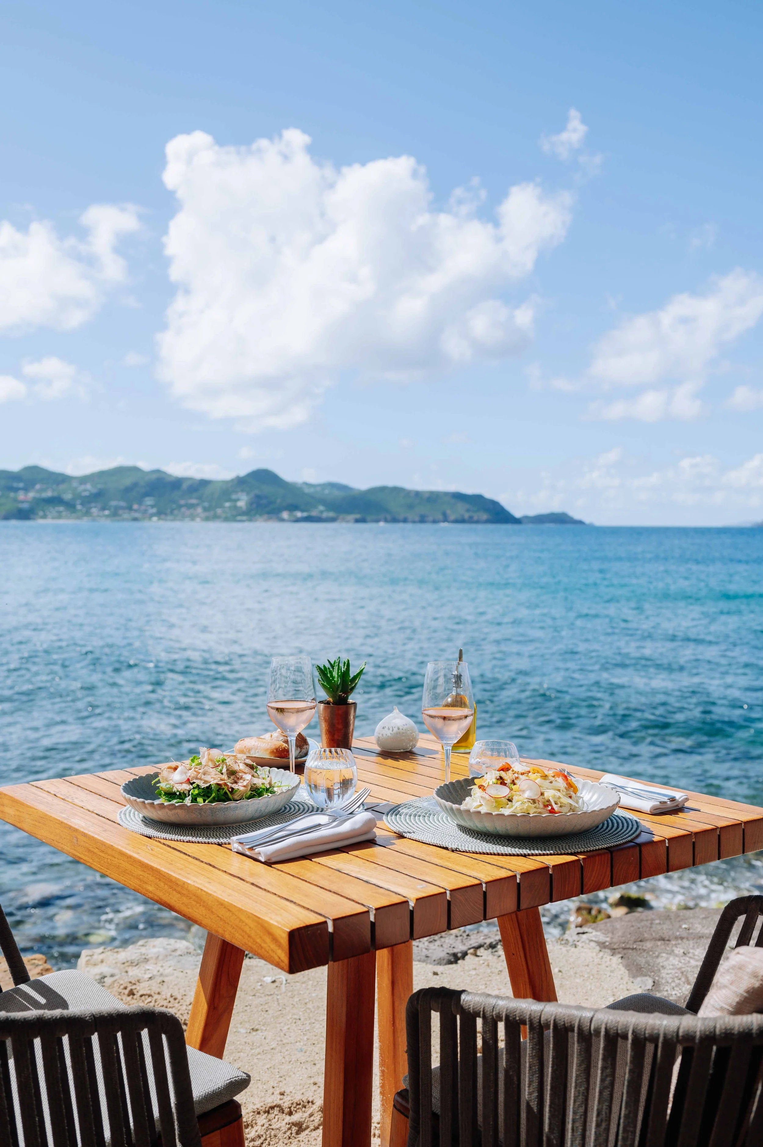 Table en bois avec deux plats de pâtes, deux verres de vin rosé, une carafe d'eau, une petite plante en pot, et des couverts, sur une plage avec vue sur la mer et des montagnes au loin, sous un ciel bleu avec quelques nuages.