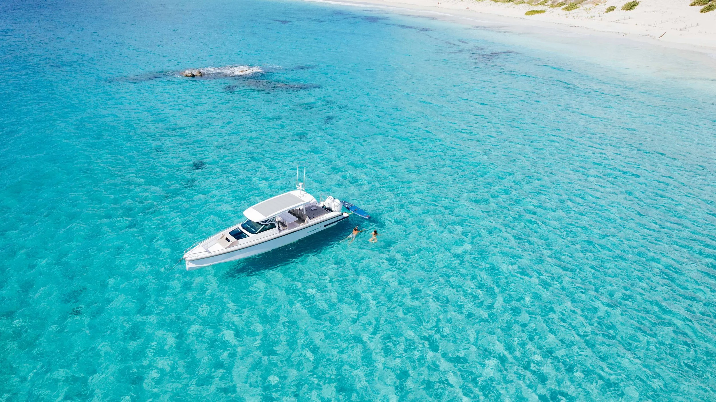 Bateau blanc flottant sur une mer turquoise claire, à proximité d'une plage de sable blanc, avec deux personnes nageant dans l'eau.