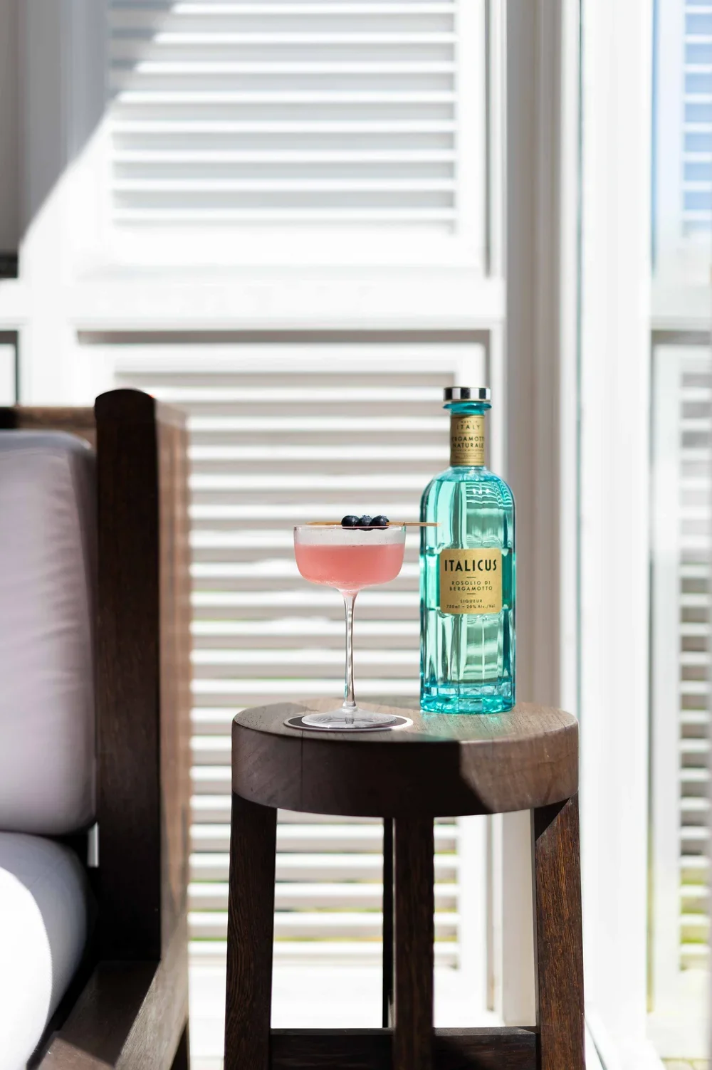 Un verre à cocktail rose avec des myrtilles, une bouteille de liqueur italienne et une fenêtre en arrière-plan profiter de la lumière naturelle.