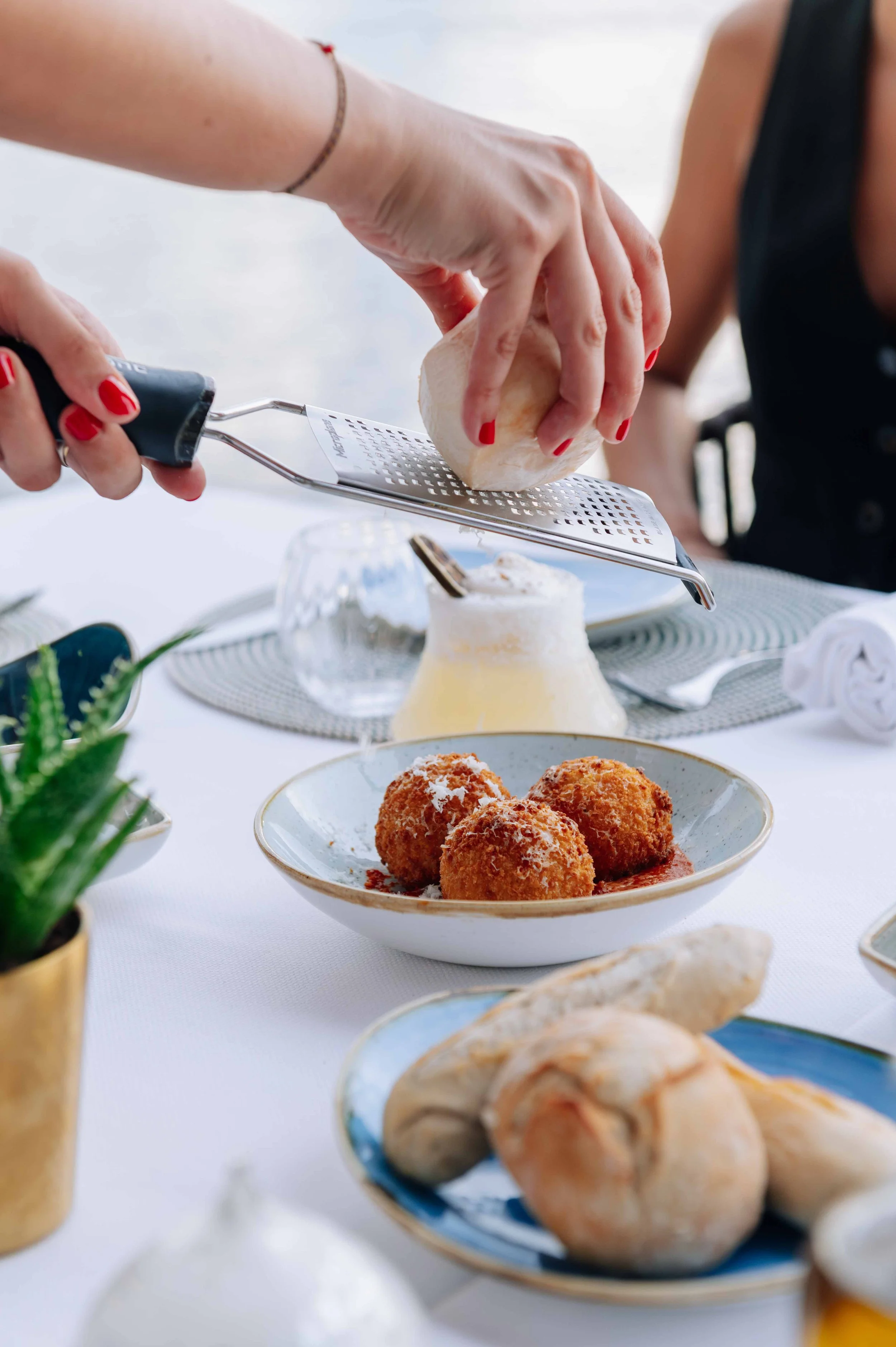 Des mains râpent du fromage sur des boulettes de viande cuites, sur une table avec divers plats et décorations.