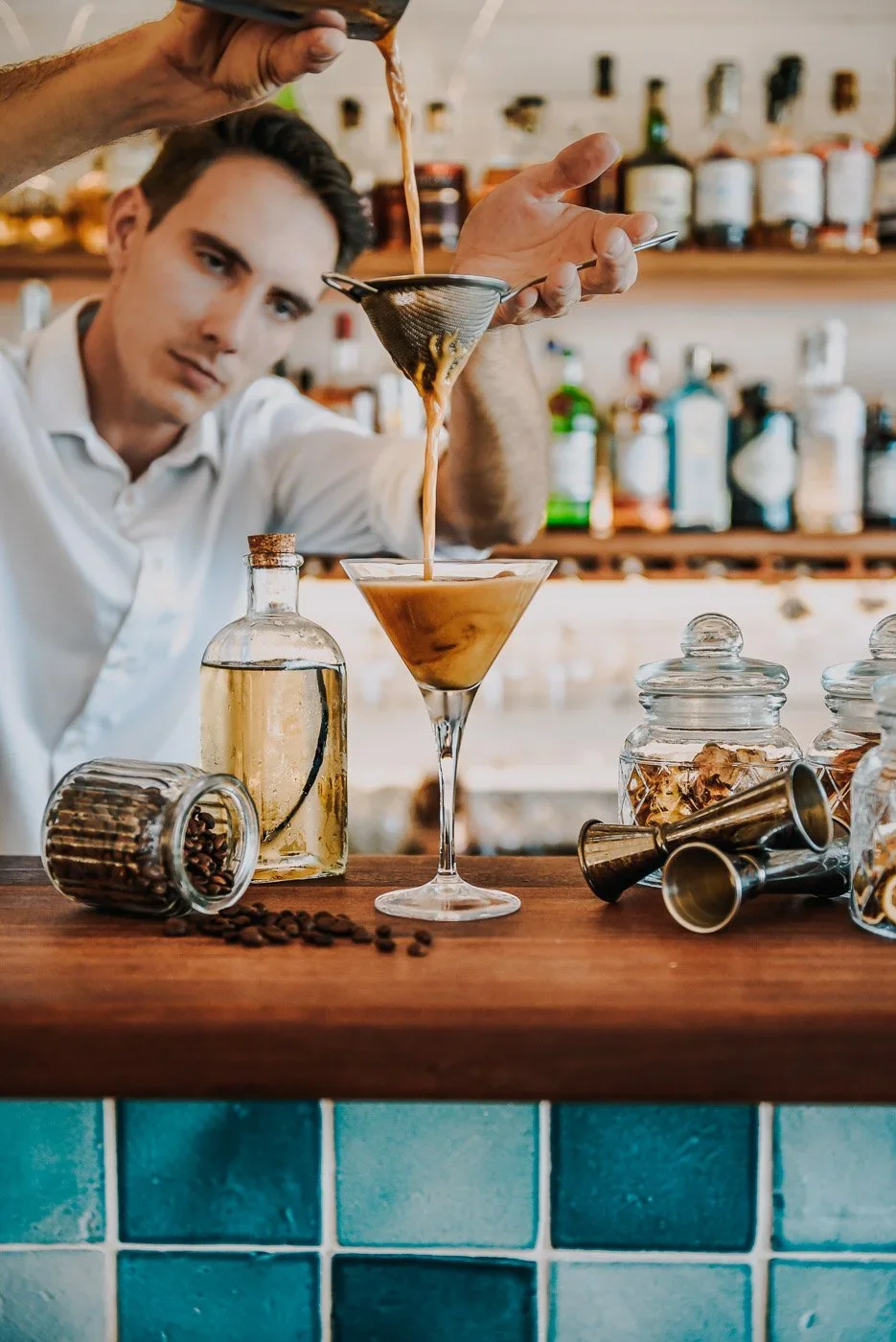 Un homme préparant un cocktail dans un bar avec des bouteilles d'alcool en arrière-plan, des bocaux, des tourets à cocktail, et des grains de café sur le comptoir en bois.