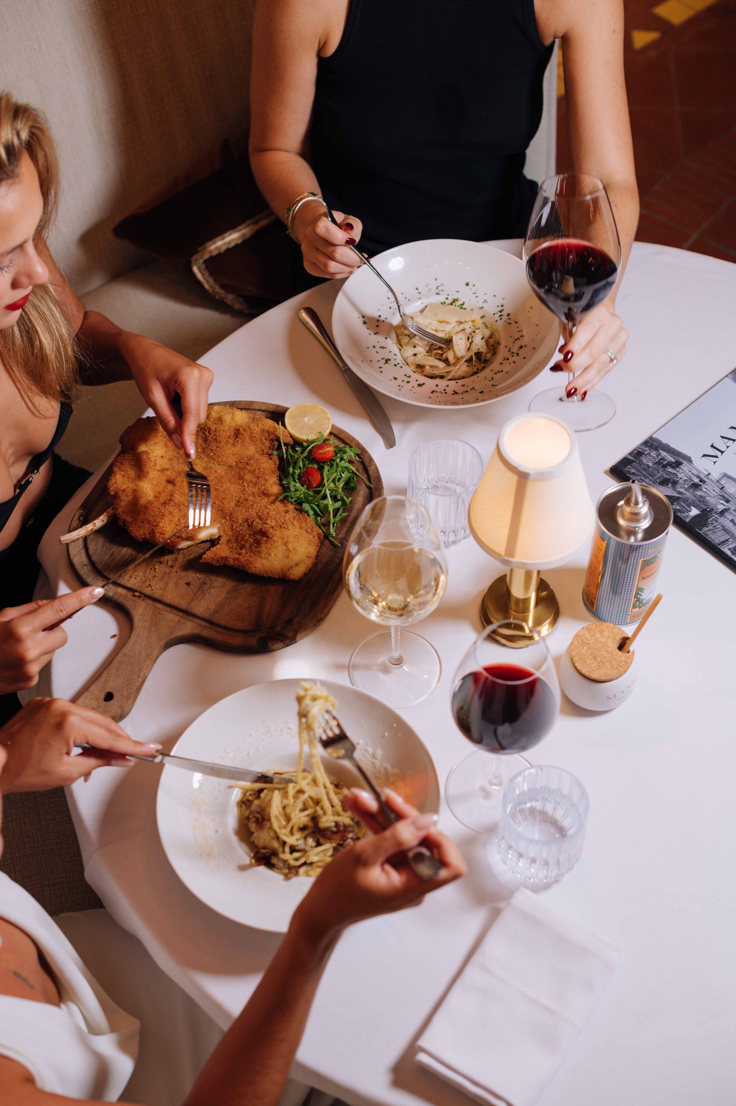 Groupe de personnes dîner autour d'une table avec plats de pâtes, escalopé de veau, pain, verre de vin rouge, verre d'eau, lampe et autres objets.