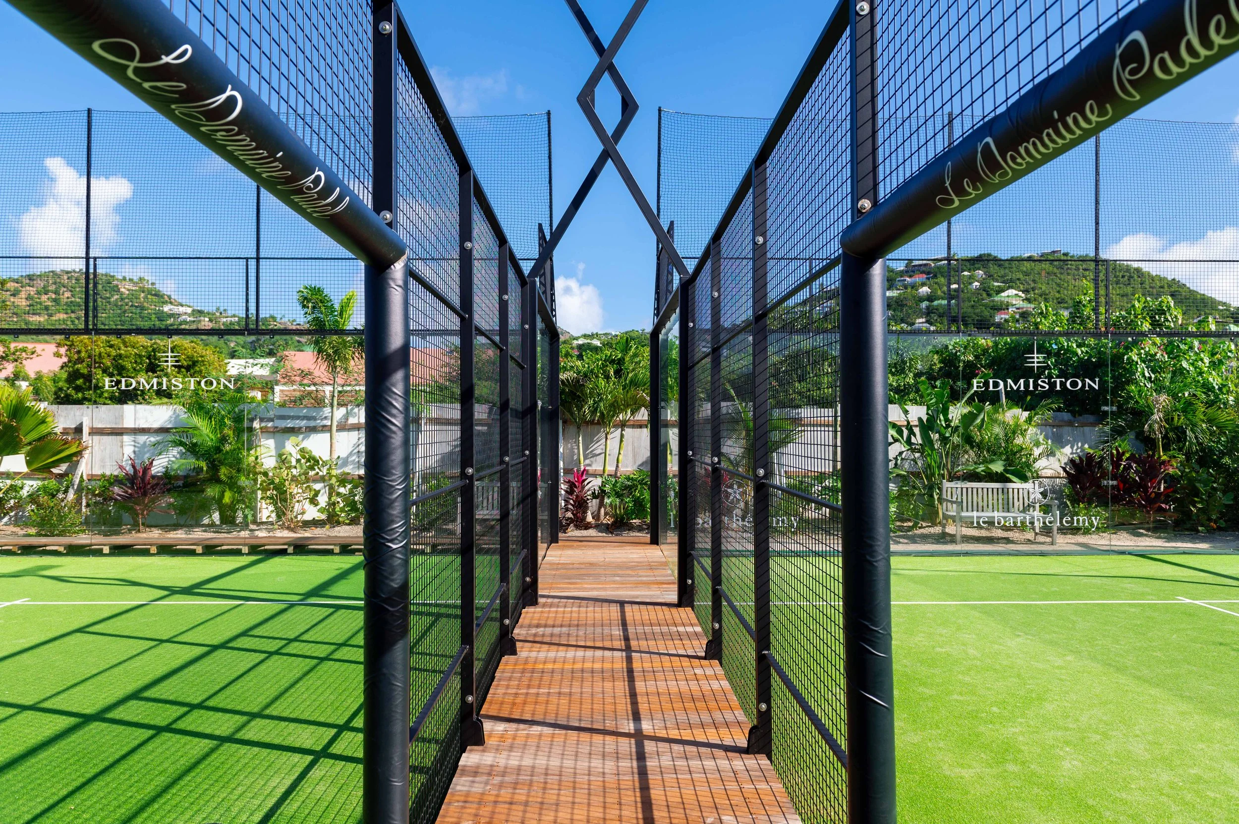 Vue d'une zone de jeu de tennis extérieure avec un portail en métal noir, des terrains en gazon synthétique vert, un décor tropical avec des arbres et des fleurs, des montagnes en arrière-plan et un ciel bleu avec quelques nuages.
