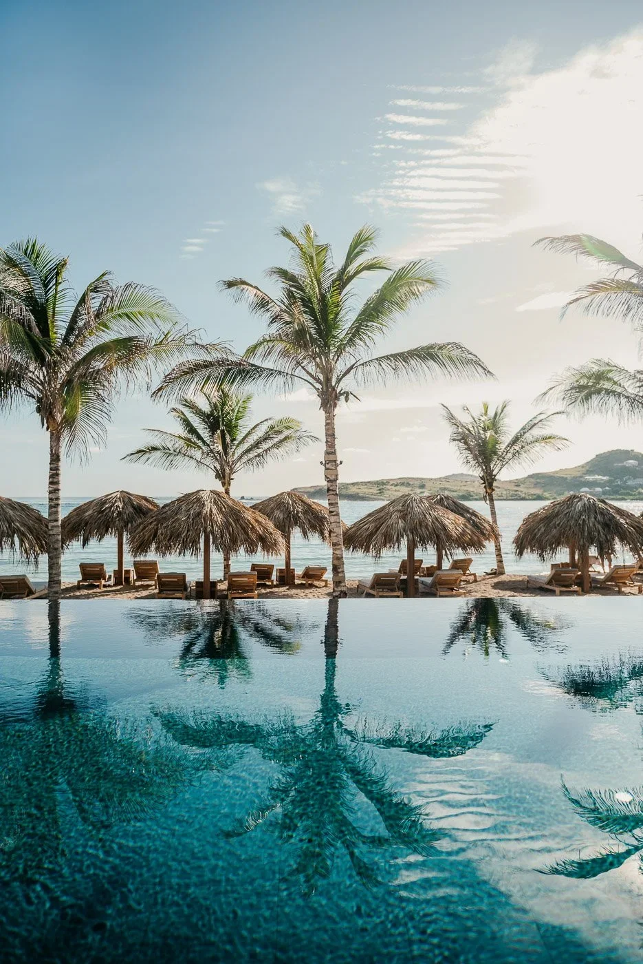 Plage tropicale avec palmiers, chaises longues sous des parasols en paille, vue sur la mer et montagnes au loin, reflet d'une palme dans une piscine au premier plan.