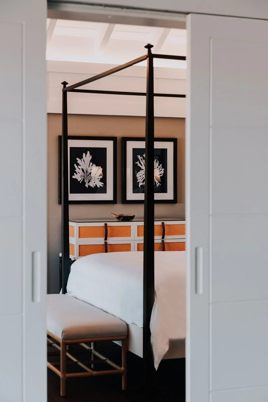 Une chambre avec un lit à baldaquin noir, des deux côtés, un chesterfield blanc avec une chaise en rotin en face du lit, et trois œuvres d'art représentant des feuilles séchées au mur