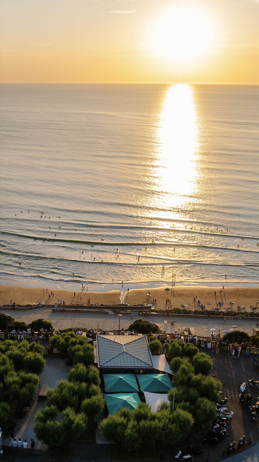 Sunset over a busy beach with people swimming and walking along the shore, viewed from a high vantage point with trees, a restaurant, and parked motorcycles in the foreground.