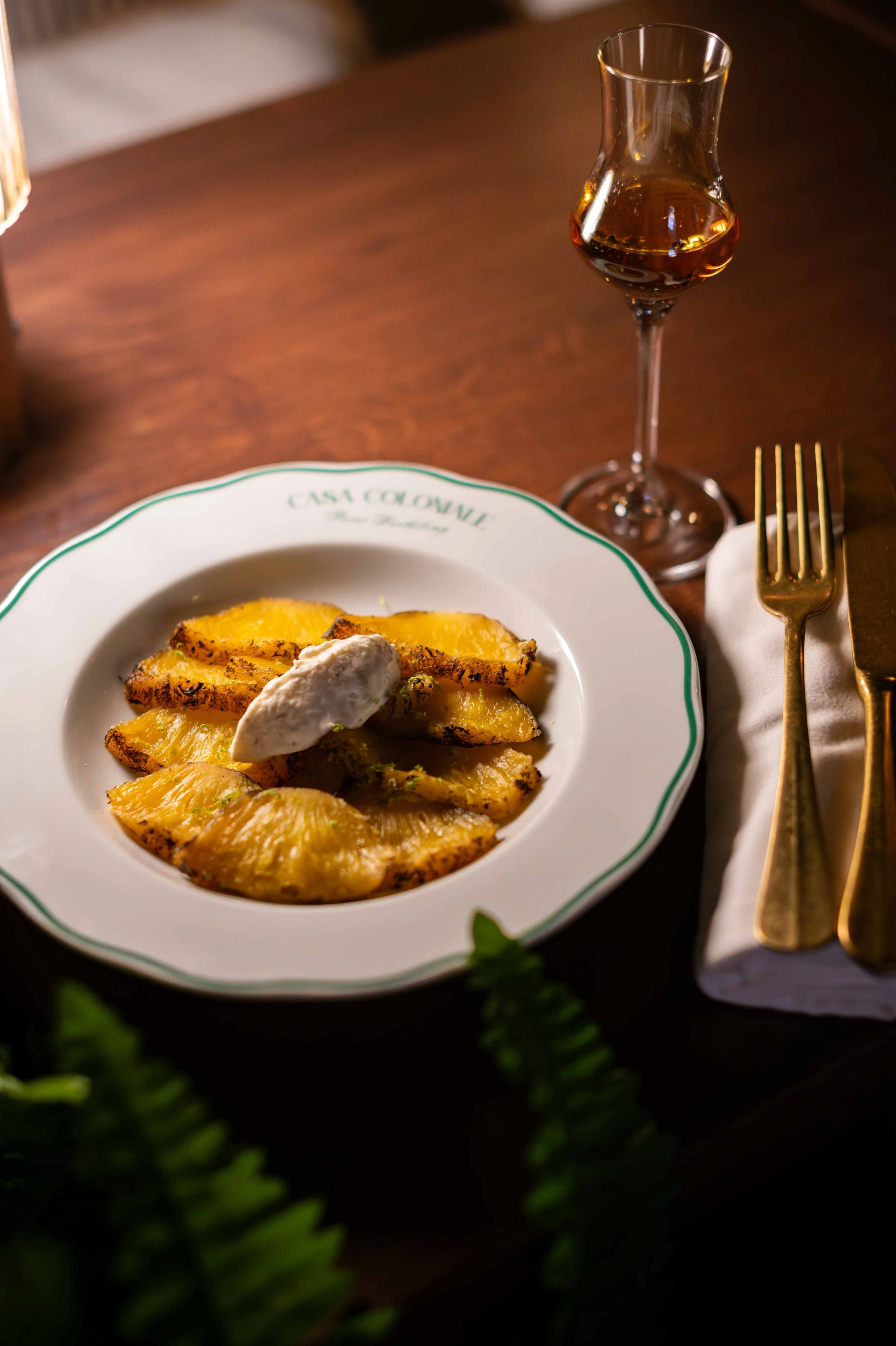 Des tranches d'ananas grillé garnies de crème ou de mascarpone, servies sur une assiette blanche avec l'inscription 'Casa Colonne'. À côté, un verre de vin ou de liqueur brun foncé, et une nappe blanche avec des couverts en or.