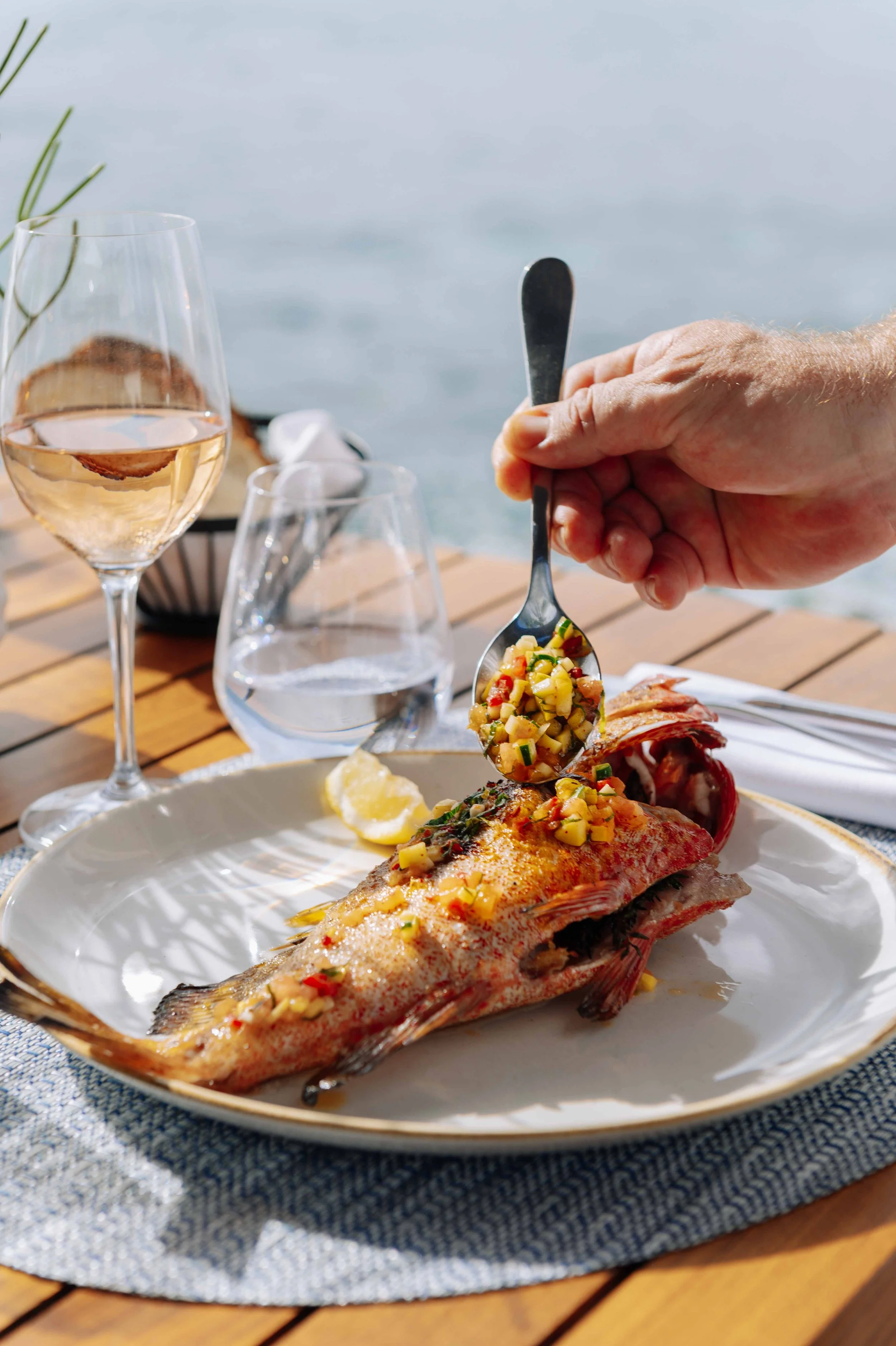 Une assiette avec un poisson frit garni d'une salsa de fruits et légumes, un verre de vin rosé, un verre d'eau, et une lumière naturelle indiquant une sortie en plein air au bord de l'eau.