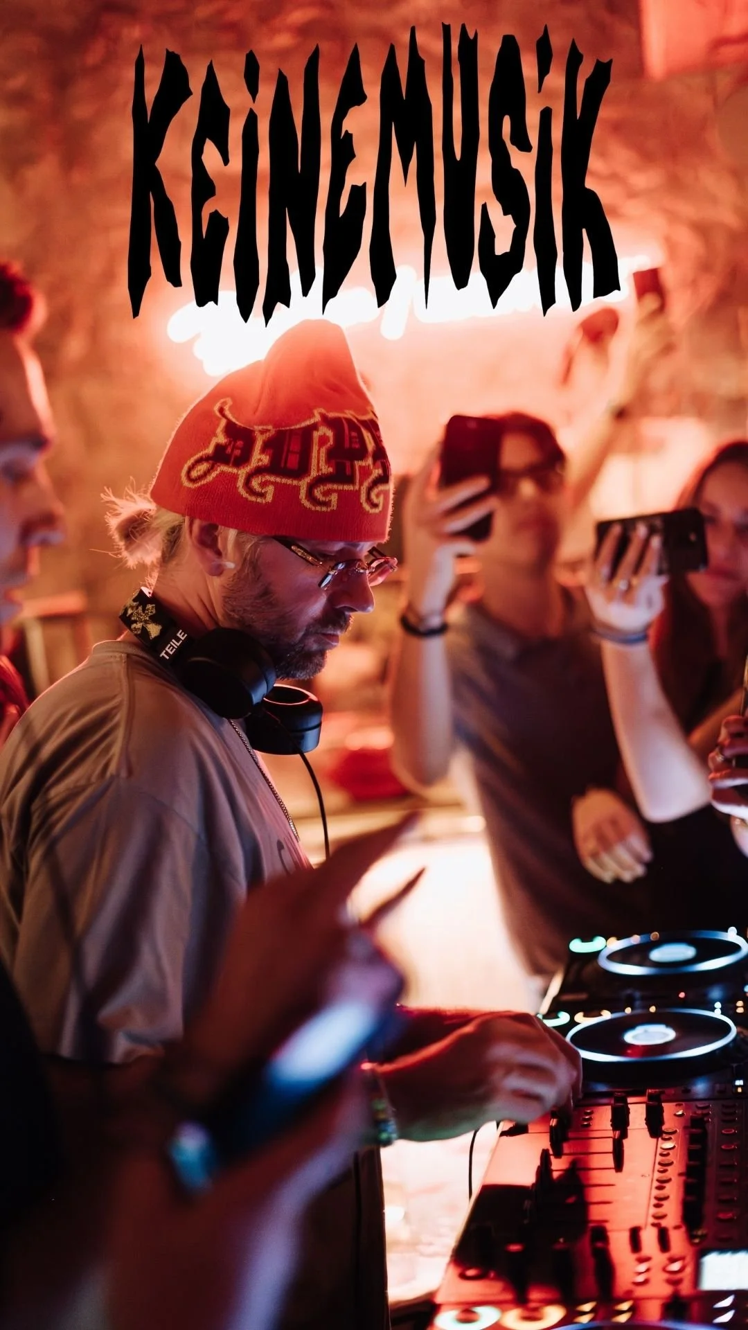 Un DJ porte un bonnet orange avec des motifs et des lunettes, jouant de la musique lors d'une fête. La scène est éclairée par des lumières chaleureuses et plusieurs personnes prennent des photos ou vidéos avec leurs téléphones.