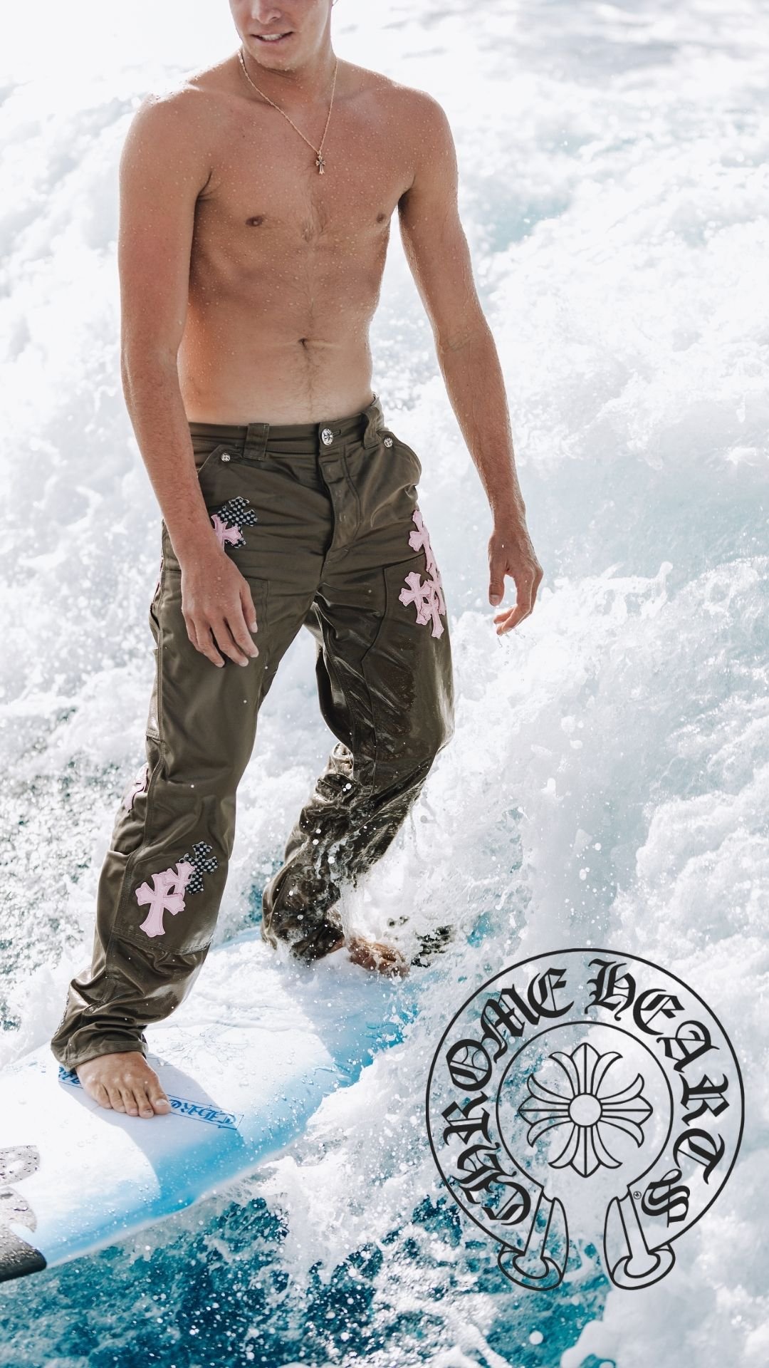 Un homme torse nu portant un collier, portant un pantalon de style cargo avec des broderies roses, qui fait du surf sur une vague dans l'océan.