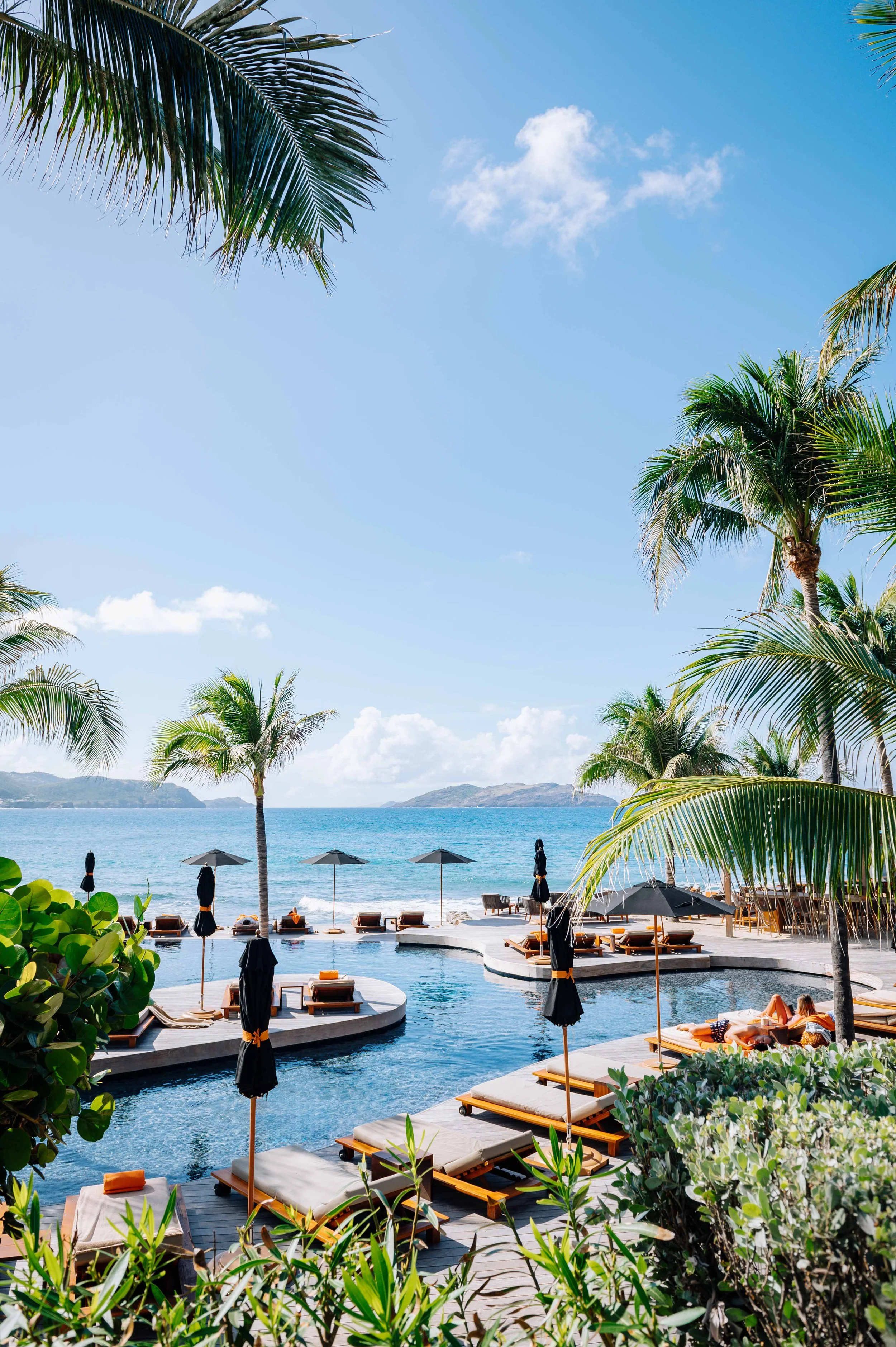 Piscine avec chaises longues et parasols à la plage, entourée de palmiers, avec la mer et des îles en arrière-plan, ciel bleu ensoleillé.