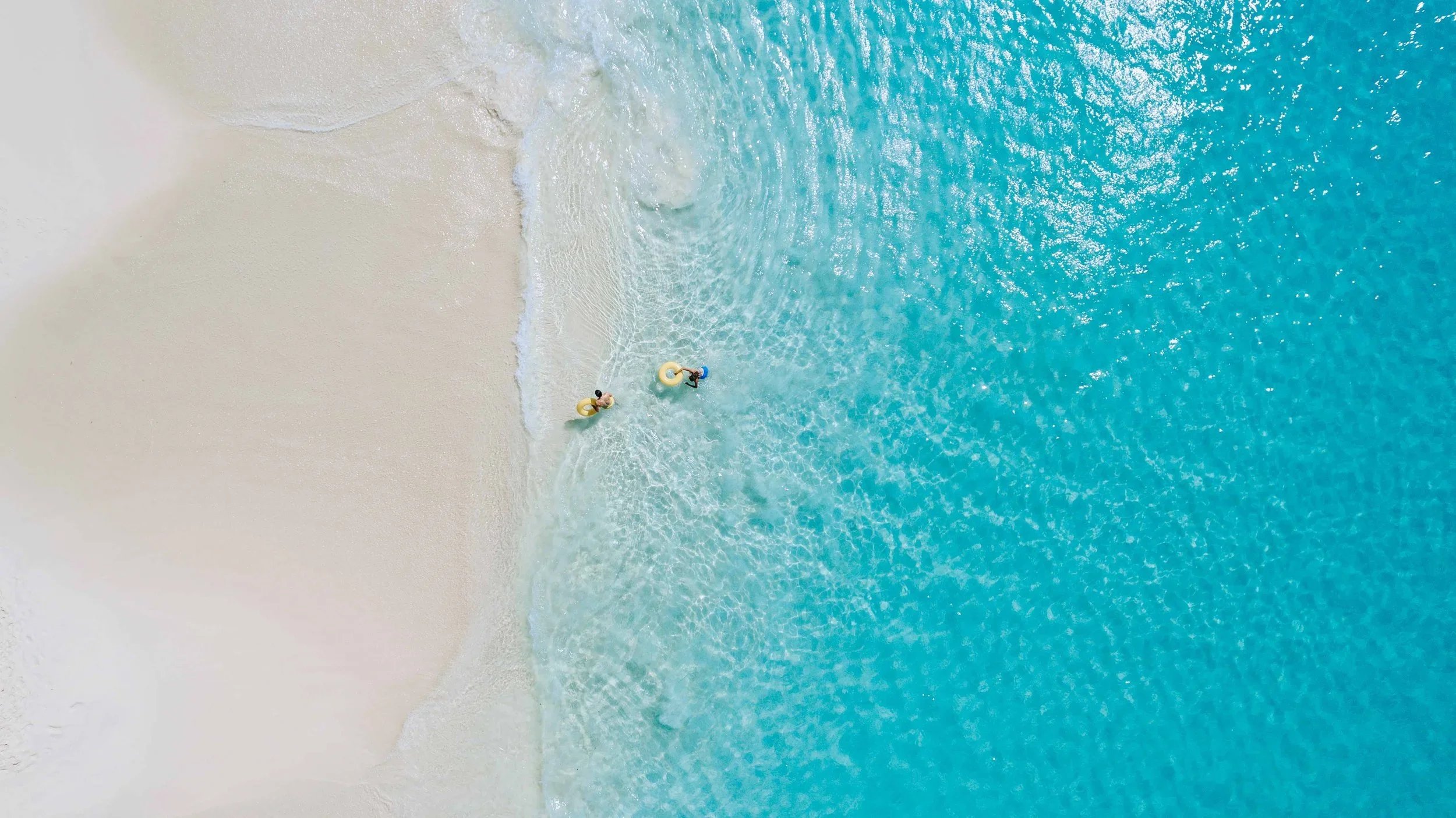 Vue aérienne d'une plage de sable blanc avec deux personnes dans l'eau turquoise, portant des flotteurs jaunes.