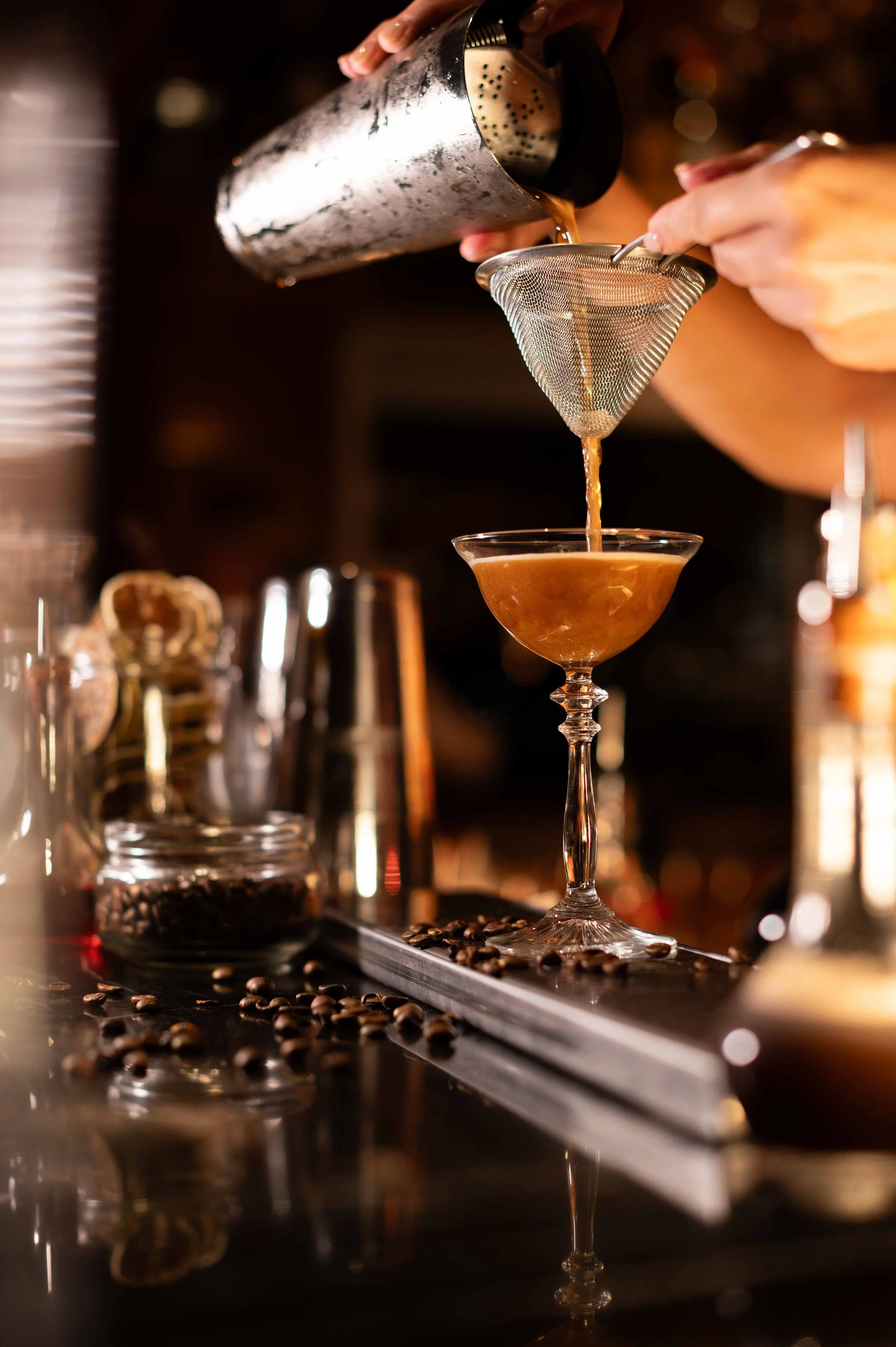 Barista verse un cocktail dans un verre élégant à travers un tamis, avec des grains de café et des accessoires de bar en arrière-plan.