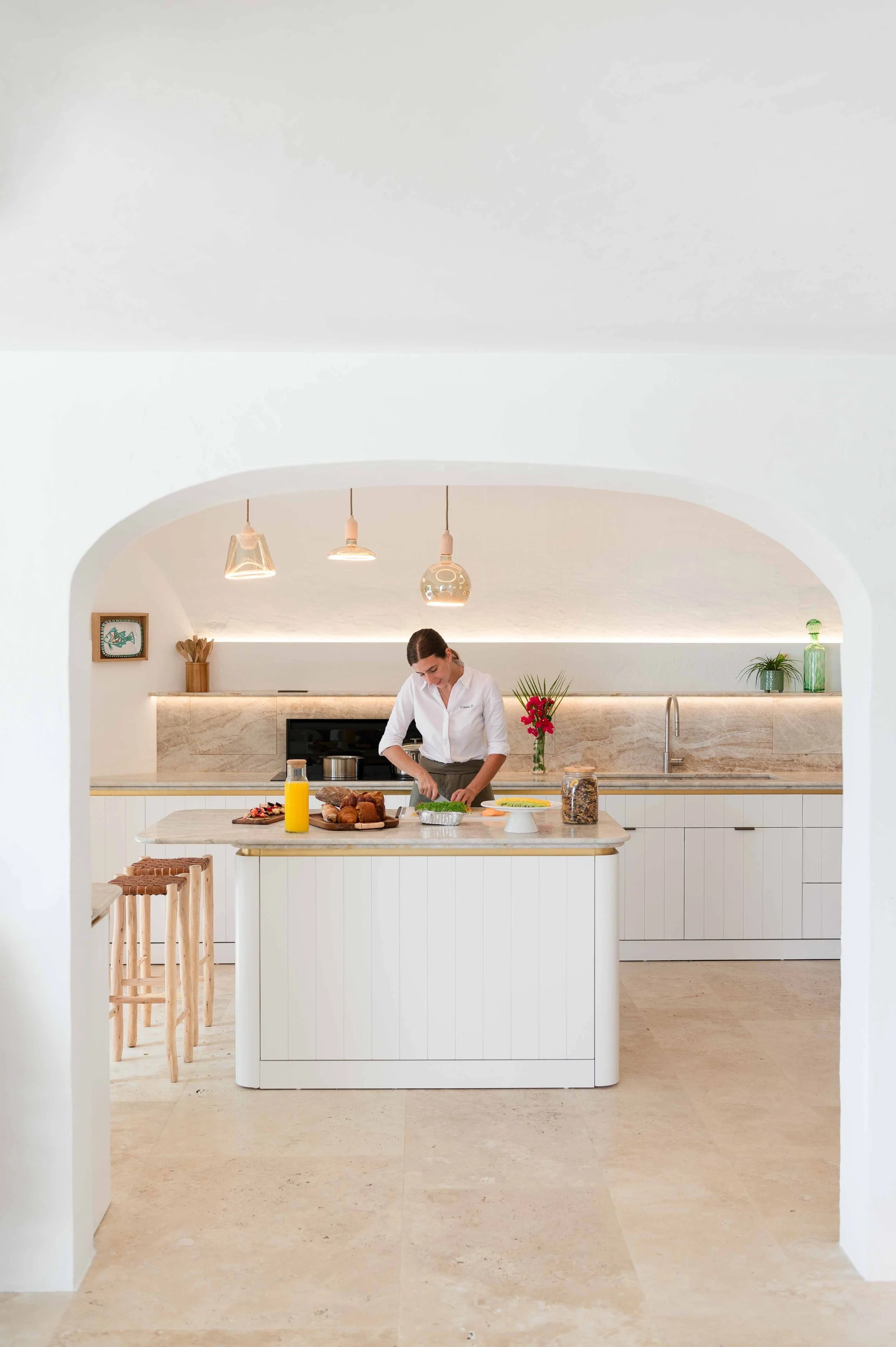 Une femme prépare un repas dans une cuisine moderne blanche avec un îlot central en marbre, plusieurs croissants, une bouteille d'huile, un bol de légumes verts et un bocal de céréales sur le plan de travail.