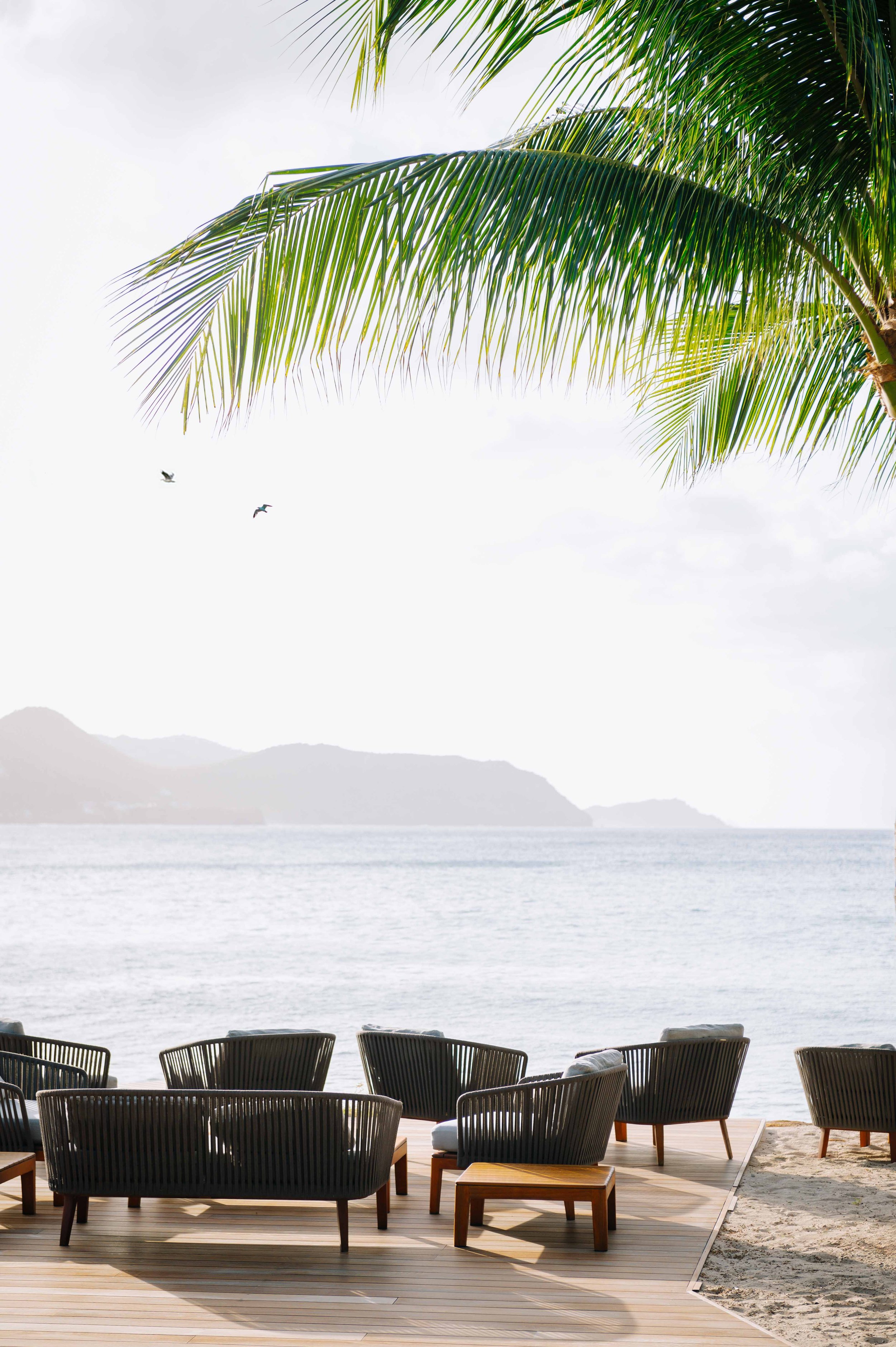 Chaises et petites tables en bois sur une terrasse en bord de mer, avec des palmiers et un ciel en arrière-plan.