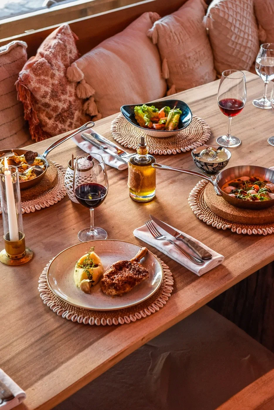 Une table dressée avec plusieurs plats, verres de vin rouge, saladier de légumes, assiette avec un morceau de viande et purée, bougies et serviettes sur un plateau en bois dans un décor cosy.