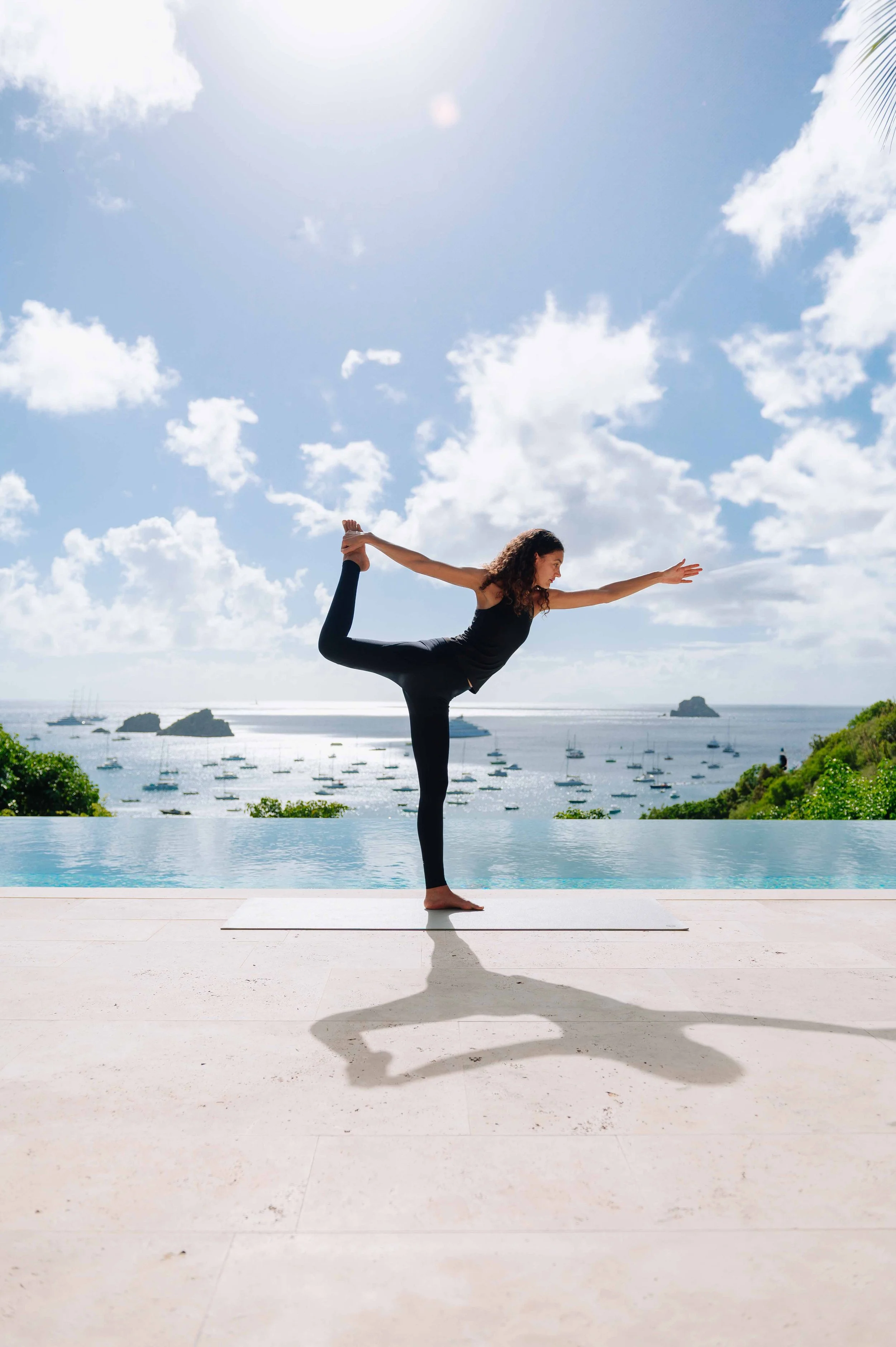 Femme faisant une posture de yoga sur une terrasse avec vue sur la mer, des bateaux et des îles en arrière-plan, ciel ensoleillé avec quelques nuages