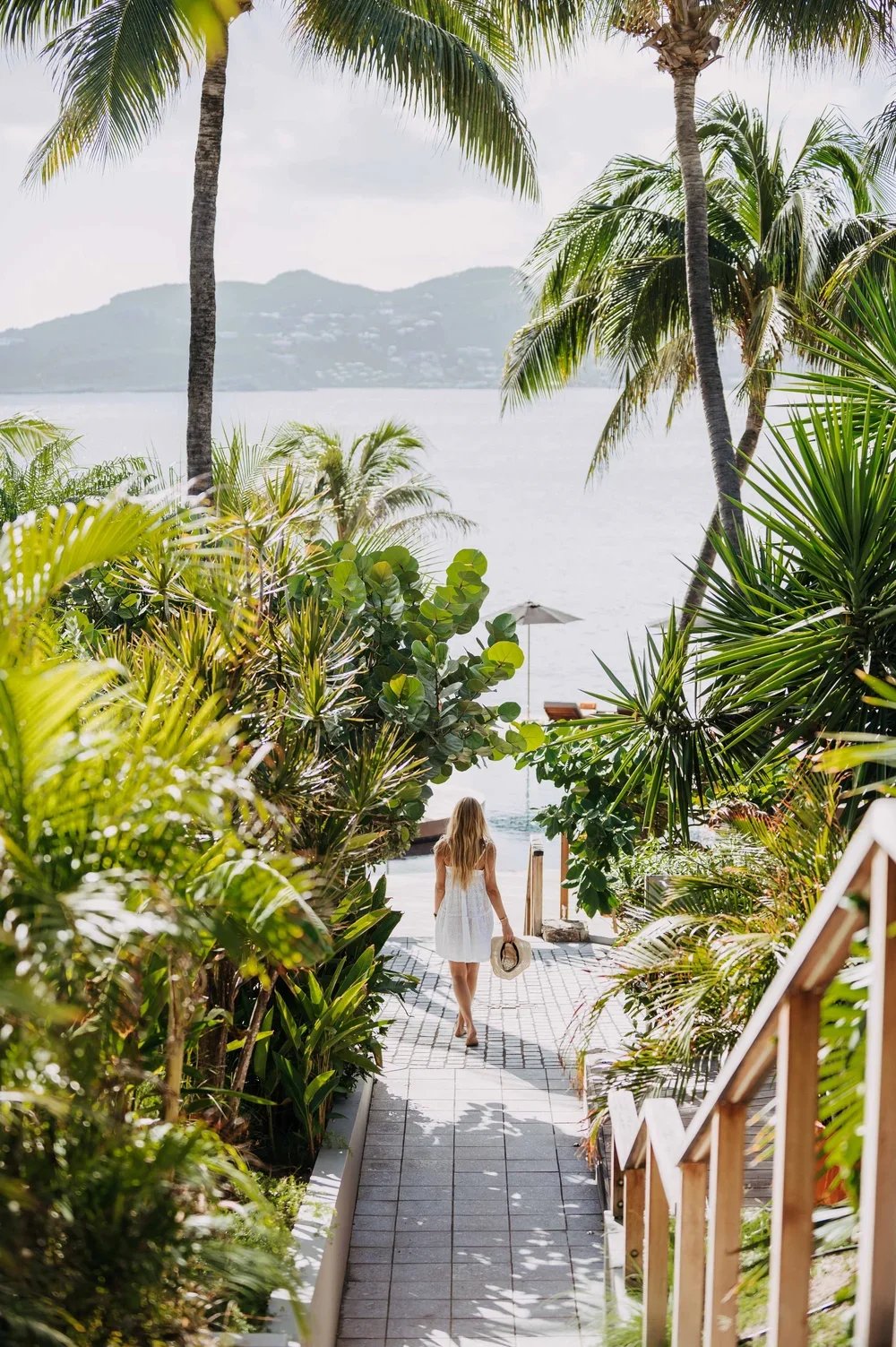 Une femme en robe blanche marche dans un chemin entouré de végétation tropicale vers une plage avec vue sur la mer et des îles au loin.
