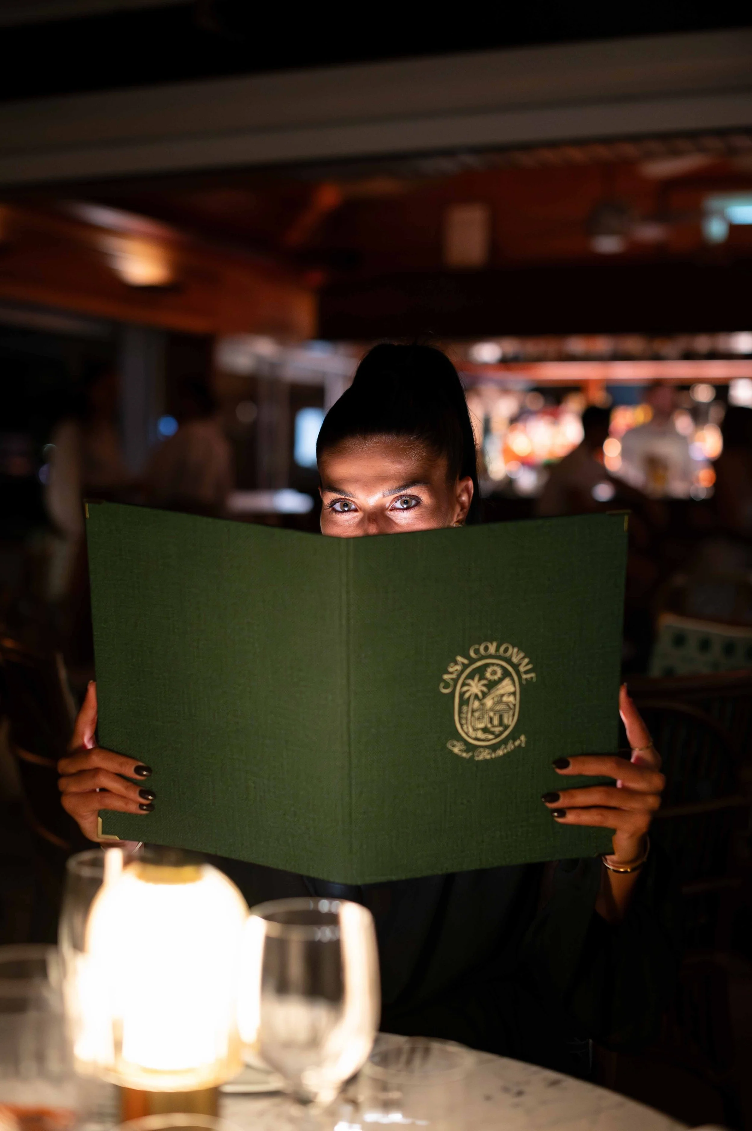 Une femme assise dans un restaurant, tenant un menu vert avec l'inscription 'Cais Colonne', regardant par-dessus le menu avec les yeux qui brillent dans une ambiance chaleureuse et tamisée.
