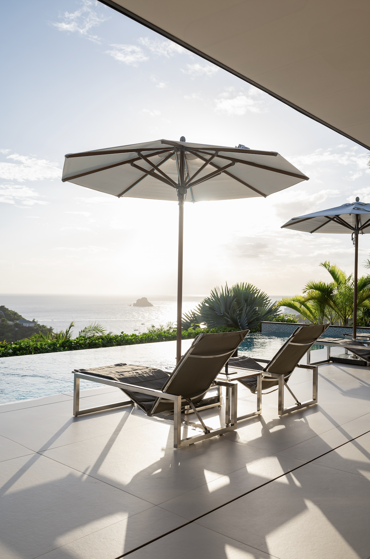 Beachside pool area with lounge chairs, umbrellas, green plants, and a view of the ocean at sunset.