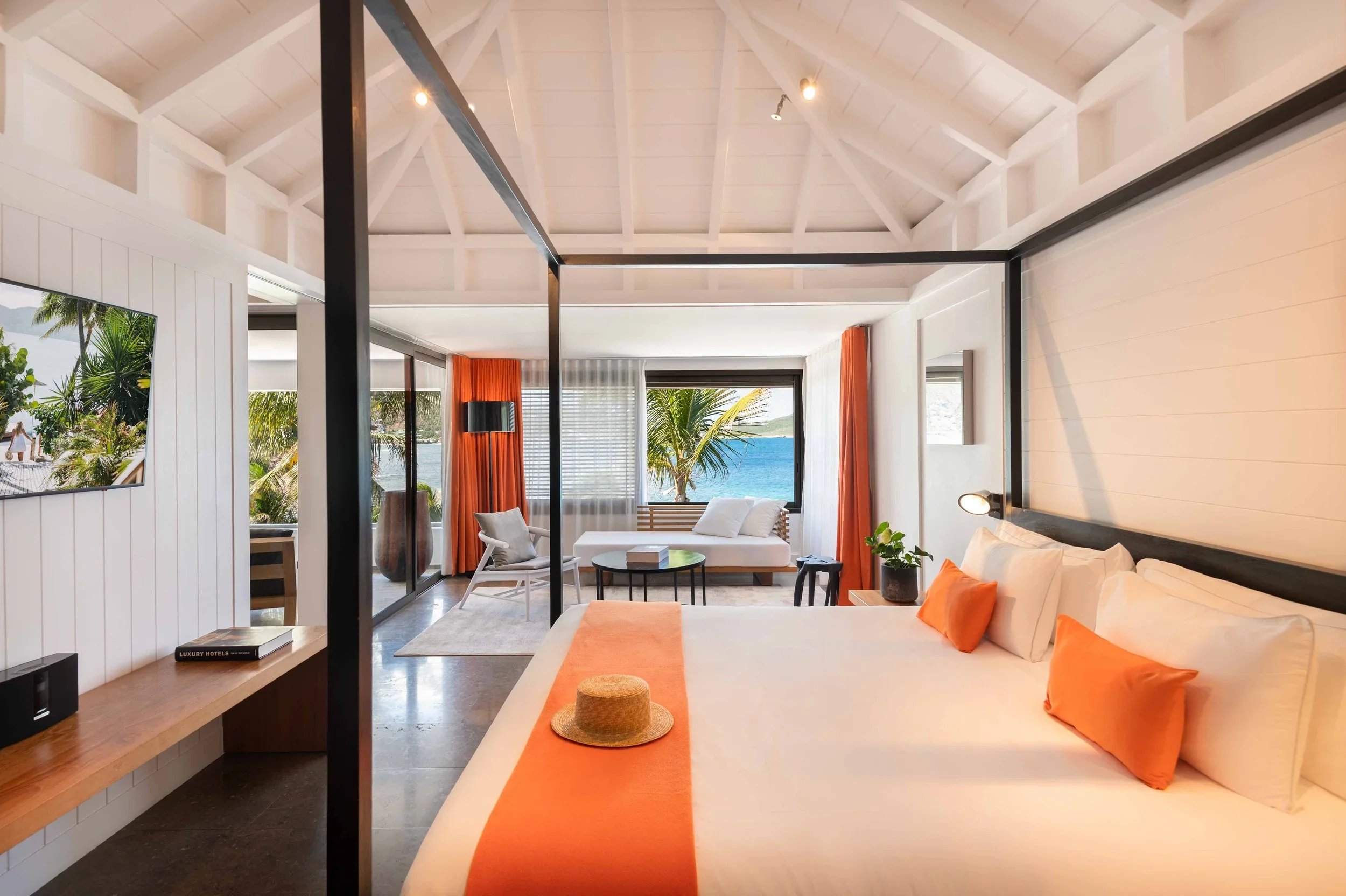 Chambre de hôtel moderne avec un lit blanc, coussins orange, vue sur la mer, canapé, télévision, et décoration tropicale.