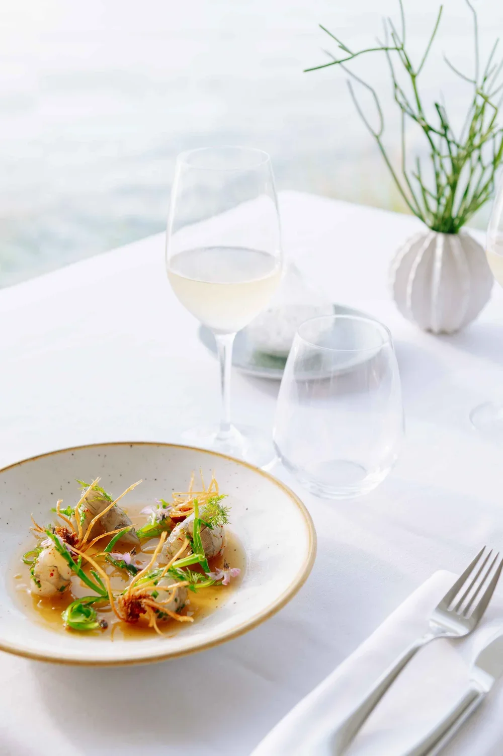Assiette de nourriture élégante avec garniture florale, banquette de table avec verre de vin blanc et verre d'eau, vase avec des plantes en arrière-plan, décor minimaliste.