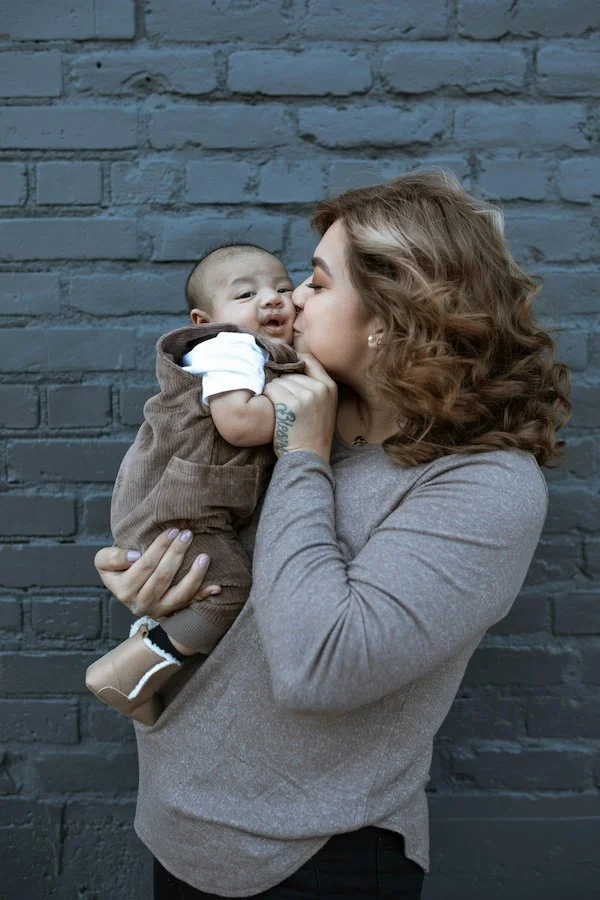 A woman with curly red hair holds a baby and kisses the baby on the cheek against a gray brick wall.