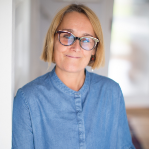 A woman with blonde hair, glasses, and a blue shirt smiling indoors.