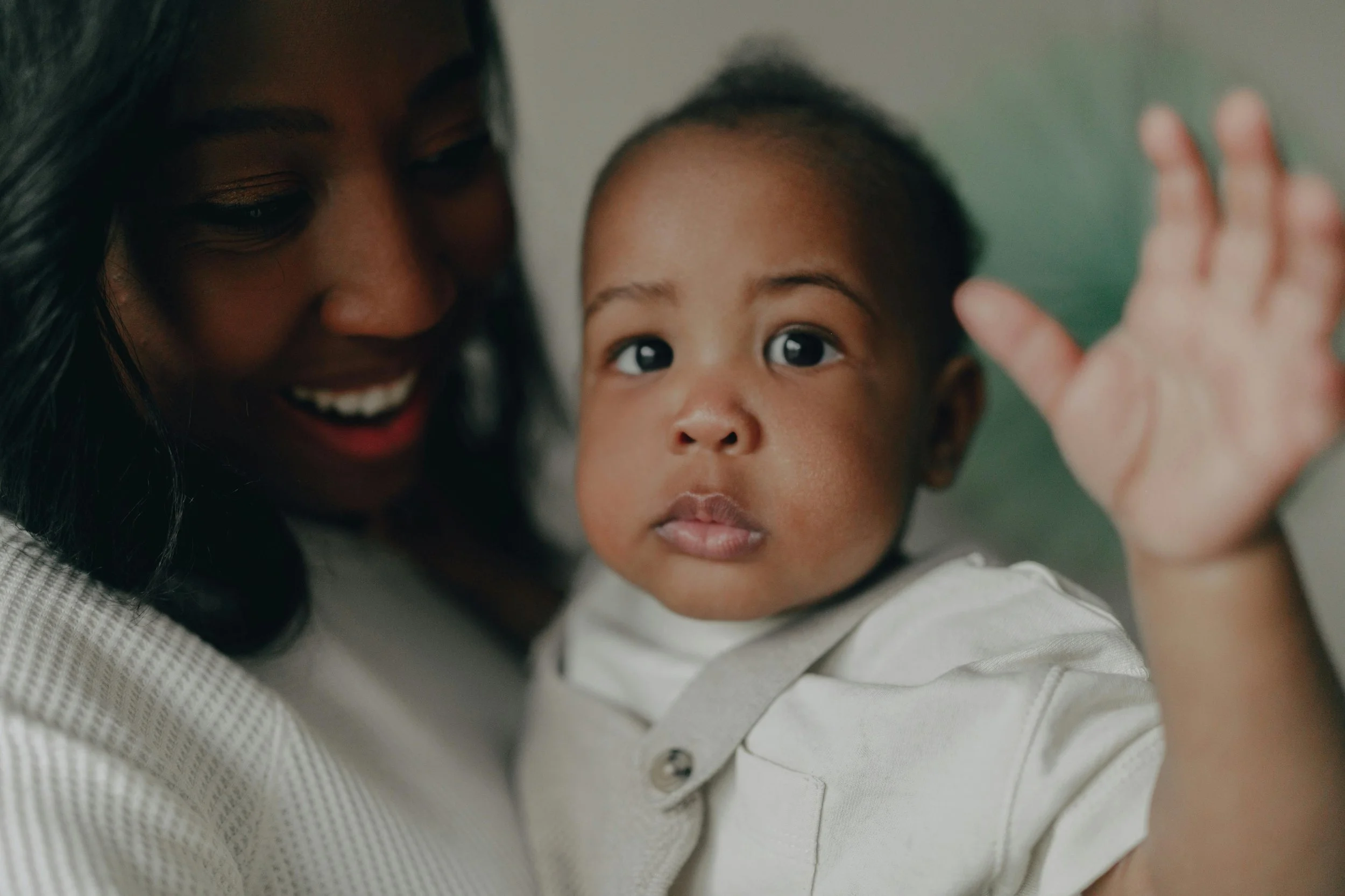 A woman smiling and holding a young child with short hair, who is looking directly at the camera with a neutral expression, while the woman is looking at the child and smiling.