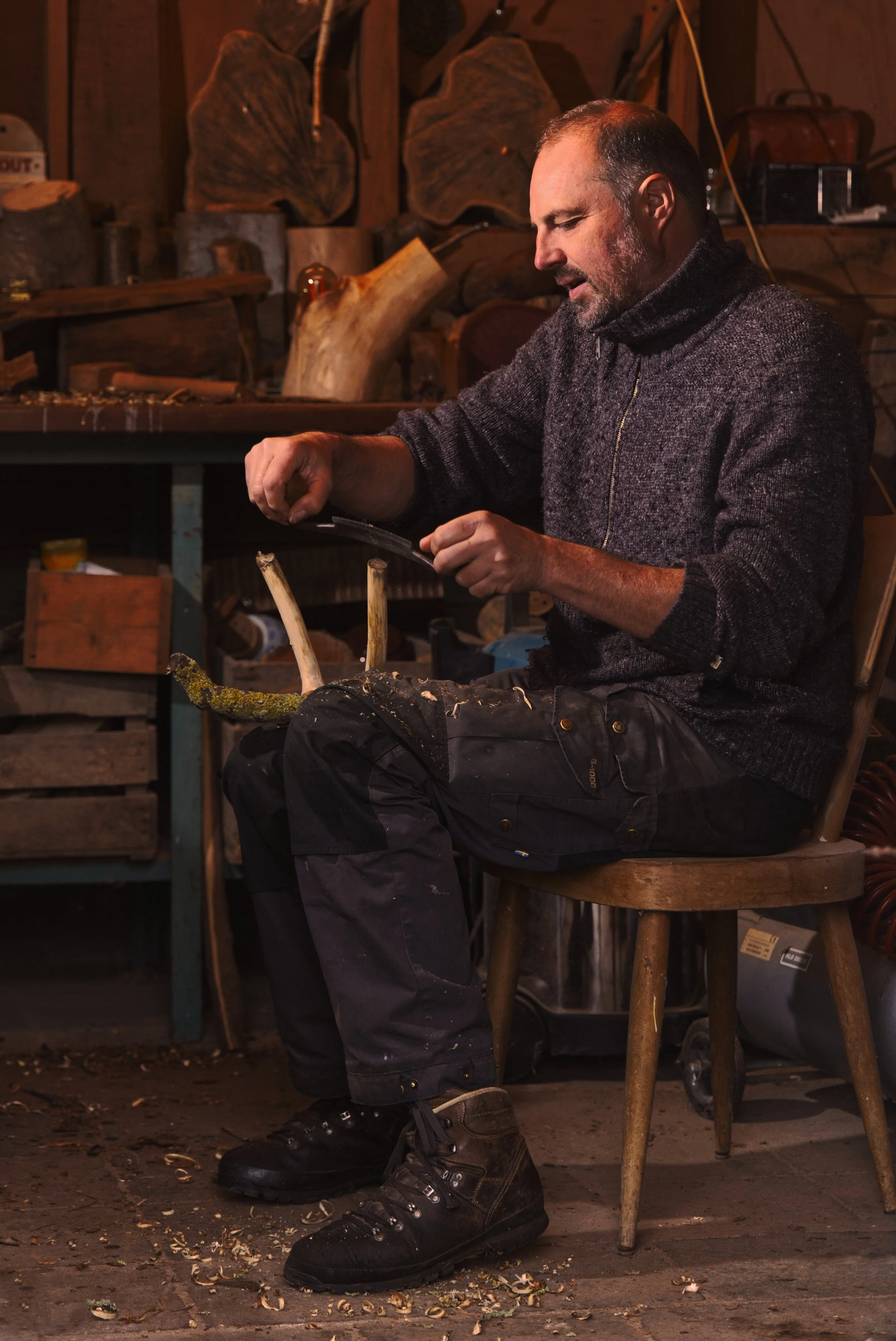 Man zit op een houten stoel in een werkplaats, werkt aan een stuk hout met een handzaag, omringd door hout en gereedschap.