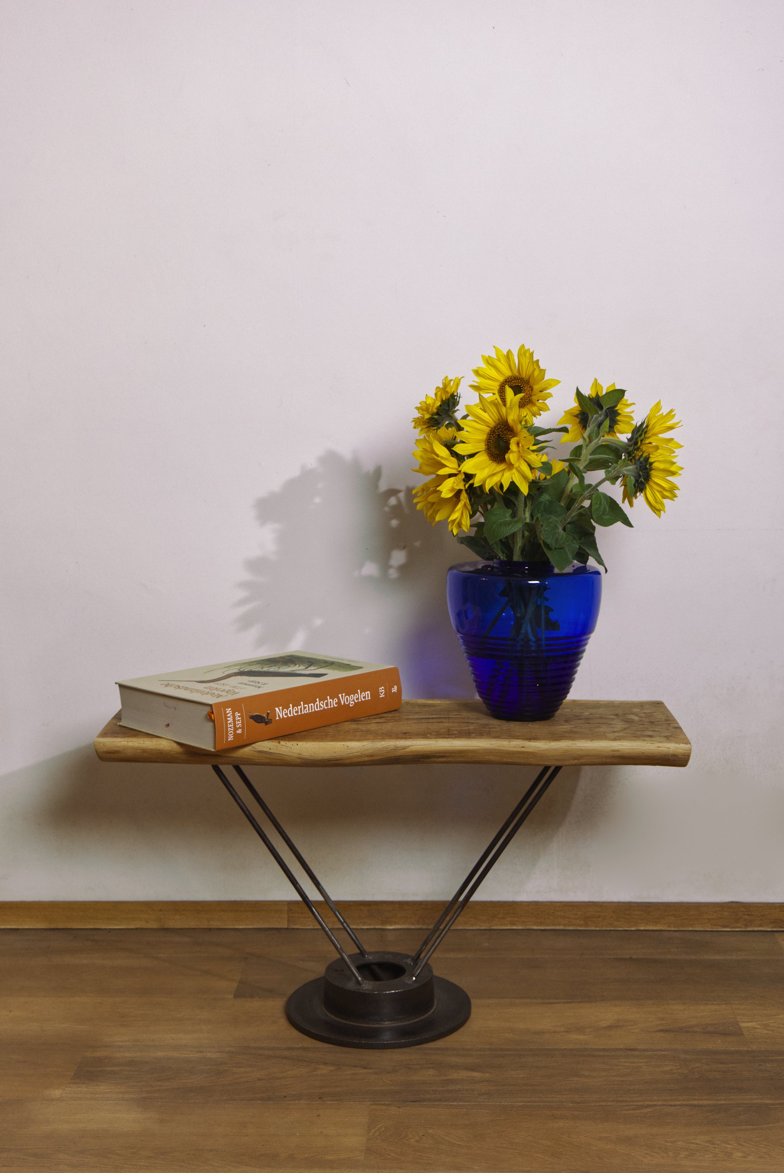 Een houten fruitschaal met metalen pootjes, een boek en een blauwe vaas met zonnebloemen